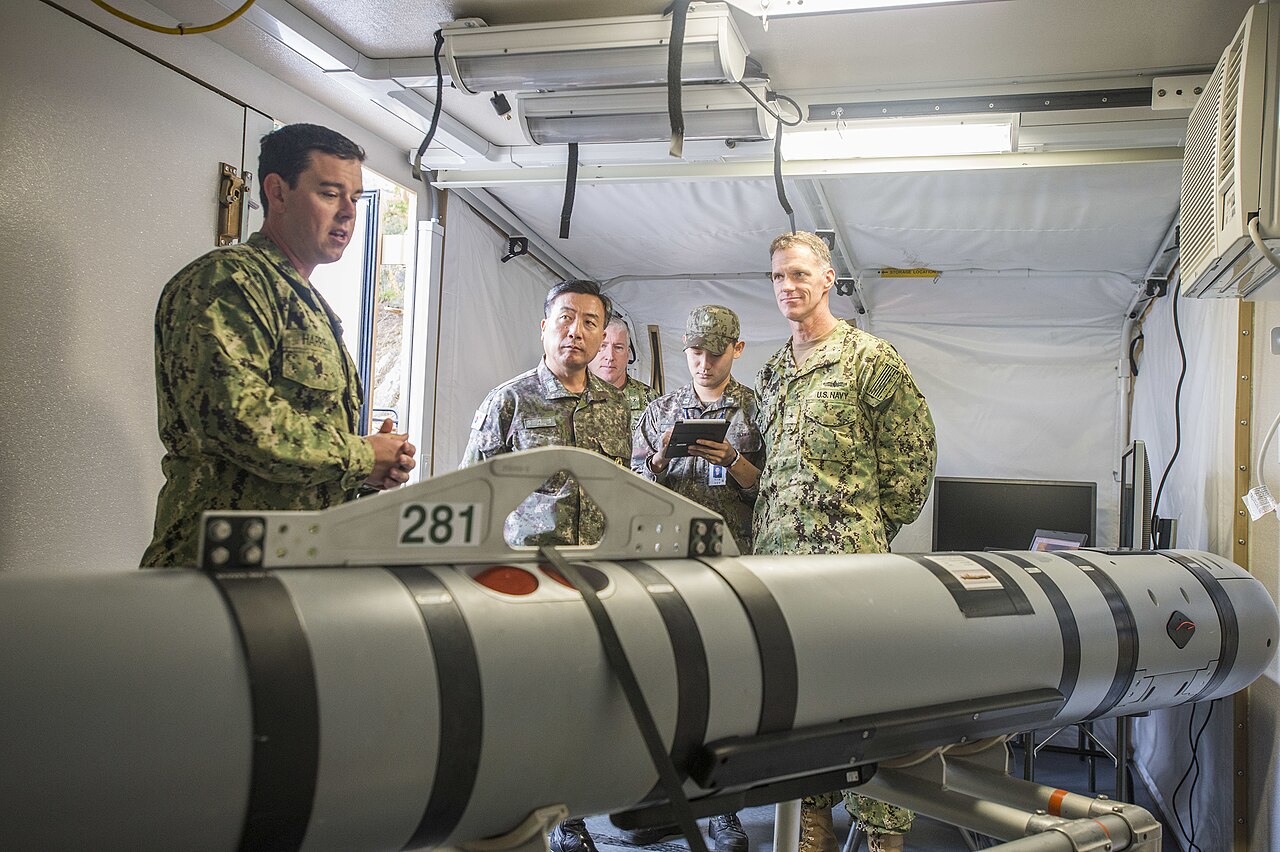 U.S. Navy sailors stand beside a long gray and black MK 18 Mod 2 Kingfish underwater drone resting on a stand indoors.