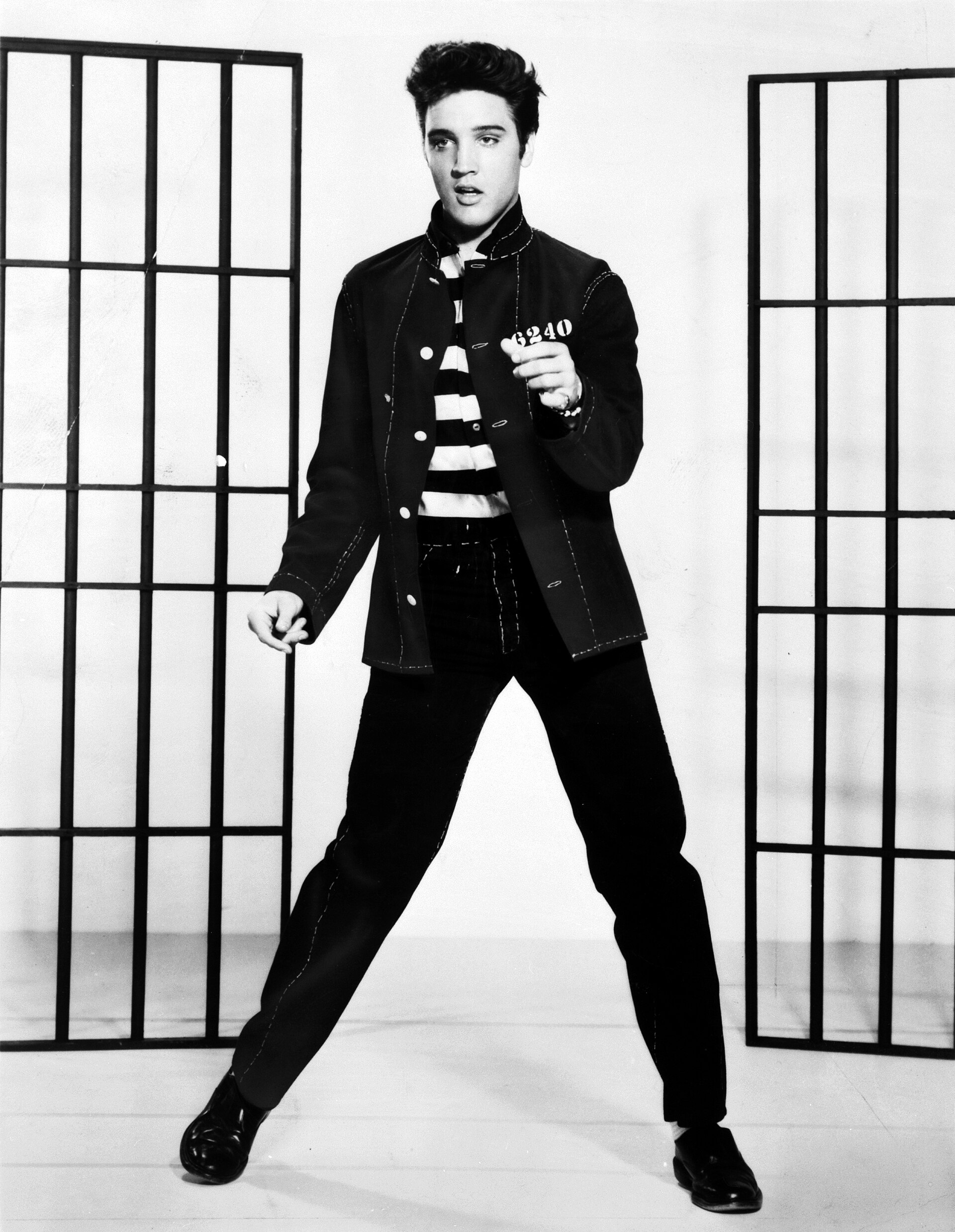 Elvis Presley, a man with dark hair, wearing a black jacket, black and white striped shirt, black pants, and black shoes, holding a card with '3240' in his right hand and standing in front of two room dividers.