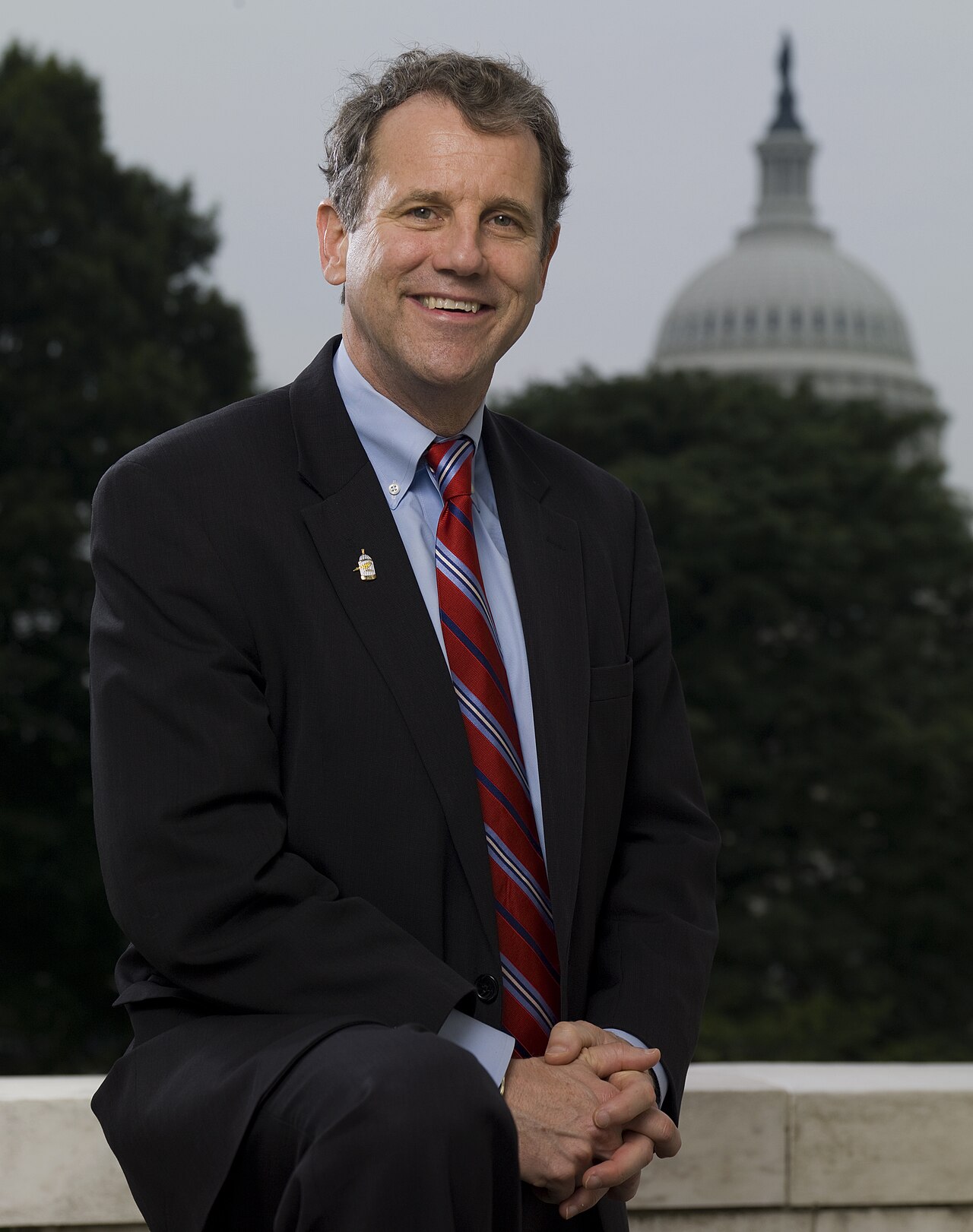 Sherrod Brown, a man in a dark suit, light blue shirt, and red and blue striped tie, smiles and clasps his hands in front of him. Behind him, out of focus, is the dome of the U.S. Capitol building.