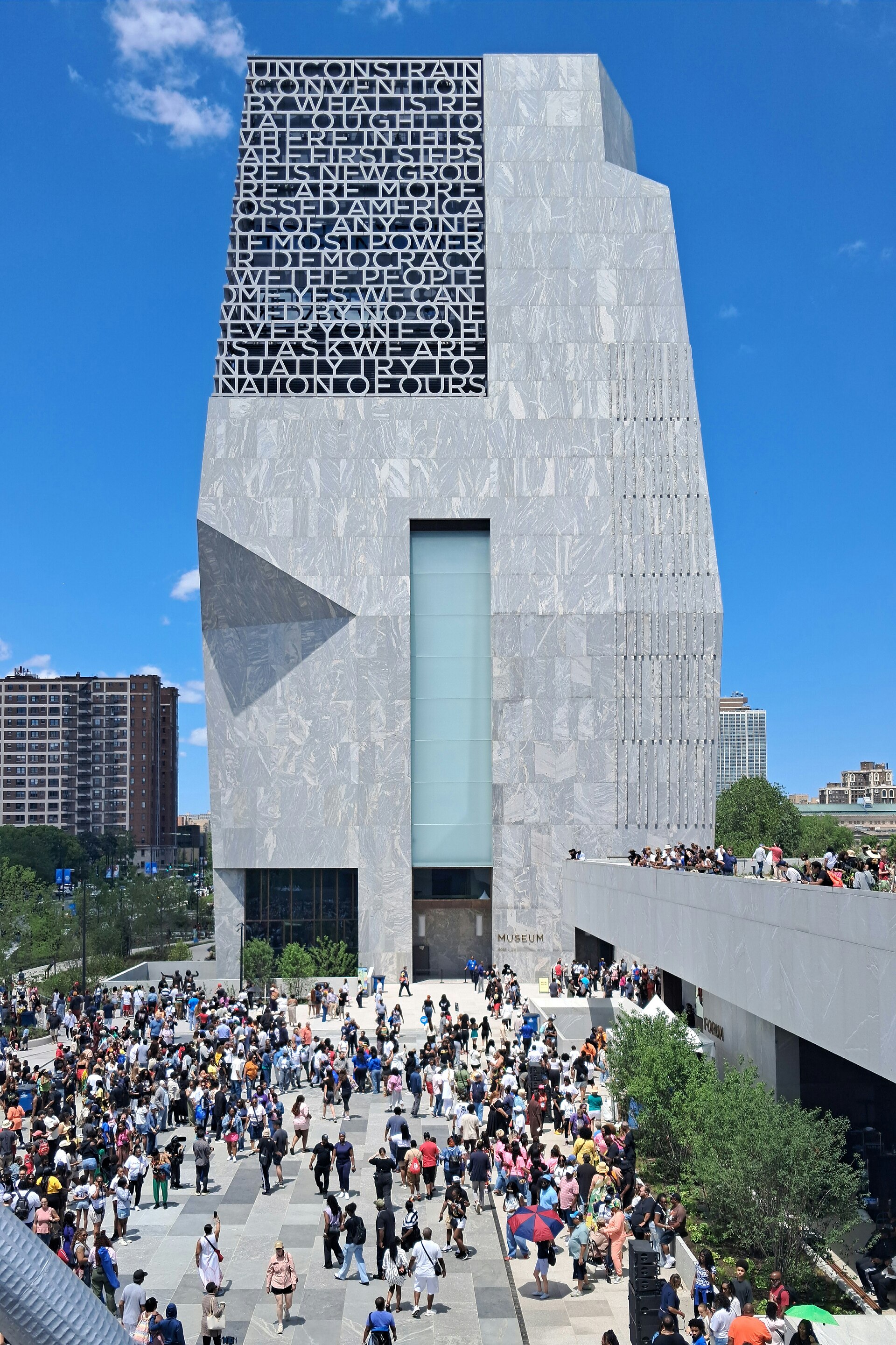 A large, modern, marble-clad building with the word MUSEUM visible on its facade, stands under a blue sky. A crowd of people are gathered in the plaza in front of the building, and on a walkway overlooking the plaza. The building has a large section with text inscribed on it.