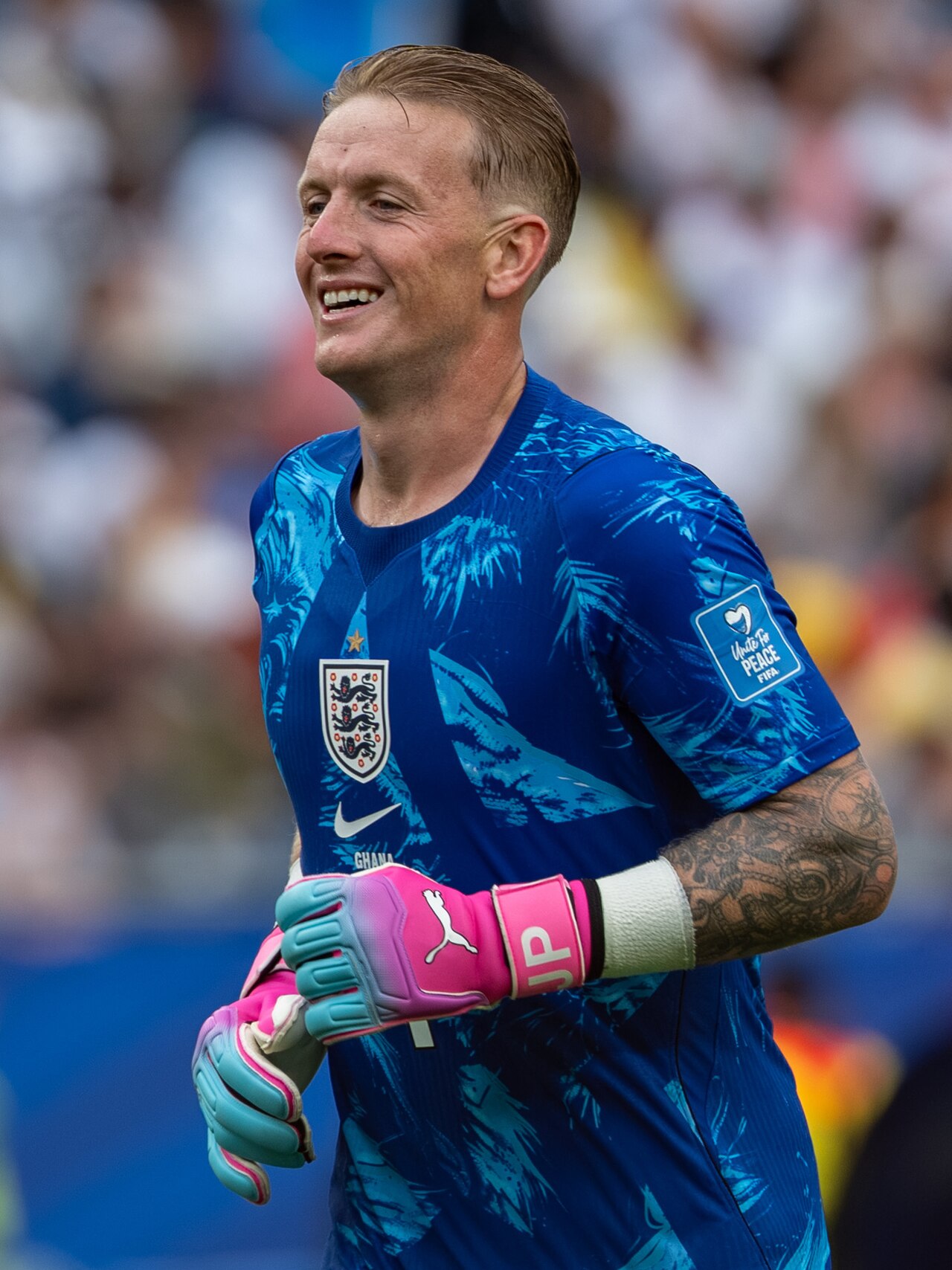 A man in a blue England football jersey with light blue accents and pink goalkeeper gloves.