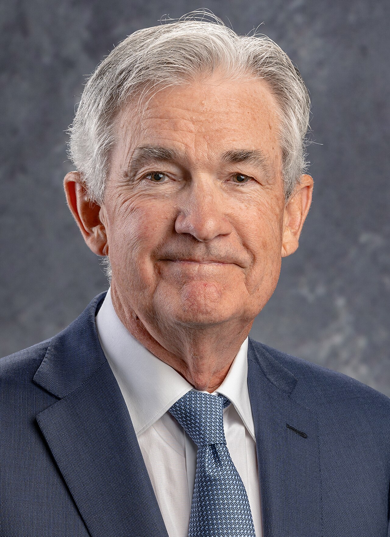 A headshot of an older man with short gray hair, wearing a blue suit, white shirt, and blue tie.