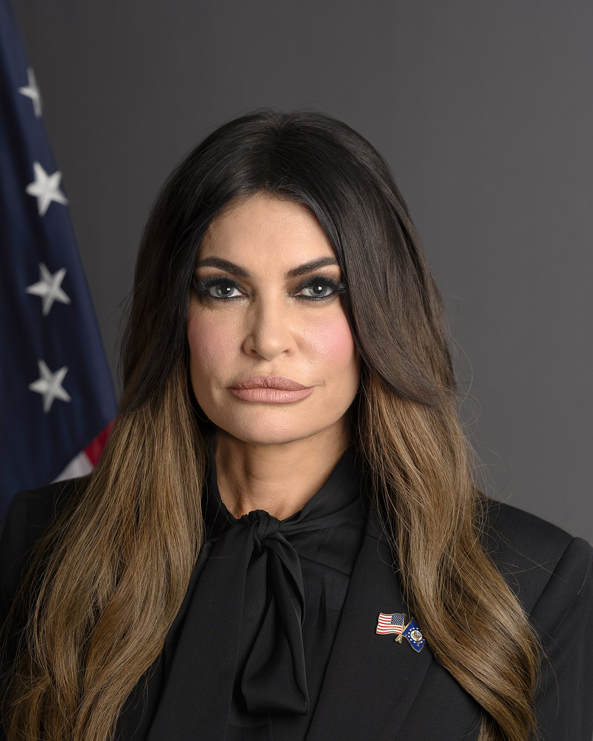 Head and shoulders of a woman with long brown hair wearing a black blouse and blazer with an American flag pin and another pin on the lapel, standing in front of a gray background with a partial American flag visible on the left.