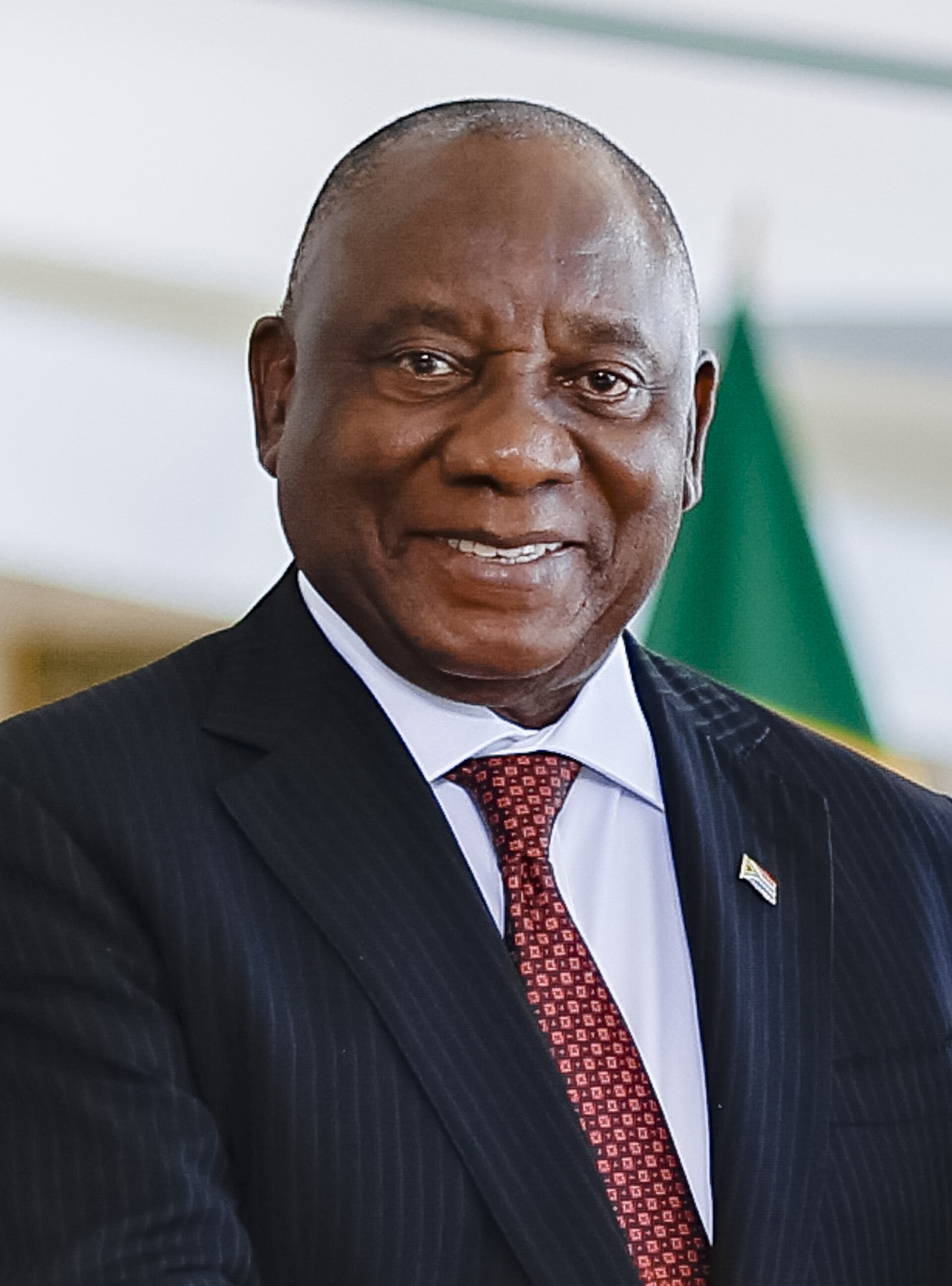 Cyril Ramaphosa, a dark-skinned man with a bald head, smiling and wearing a black suit, white shirt, and red tie with a small pin on his left lapel.