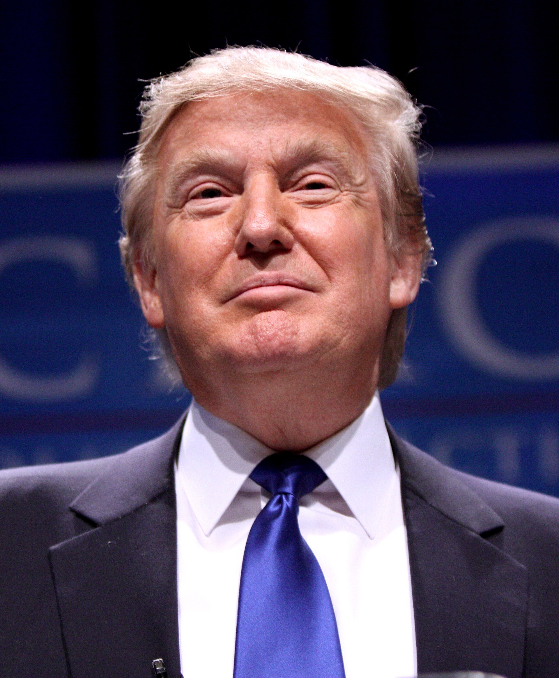 A man with short blond hair wearing a dark suit, white shirt, and blue tie