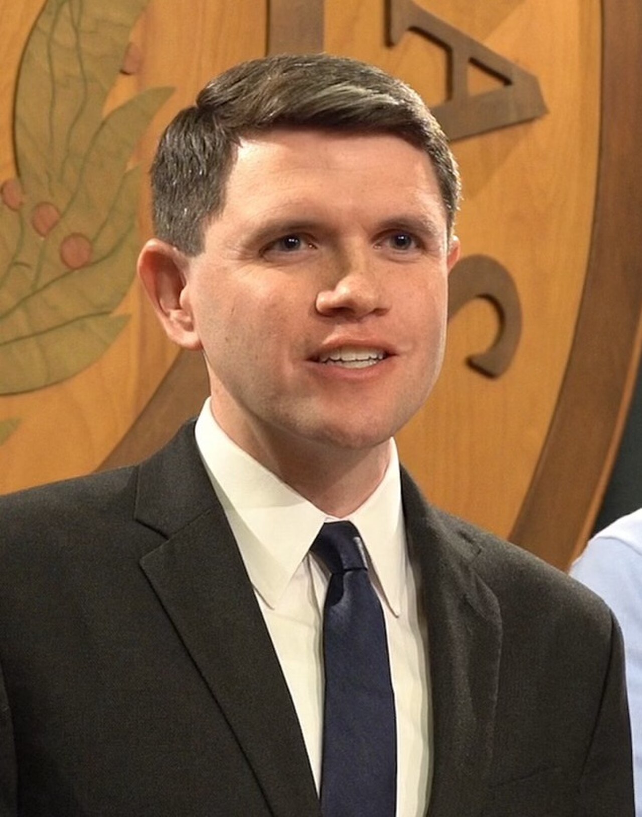 A man in a black suit, white shirt, and blue tie smiles slightly, with a wooden emblem featuring leaves and symbols in the background.