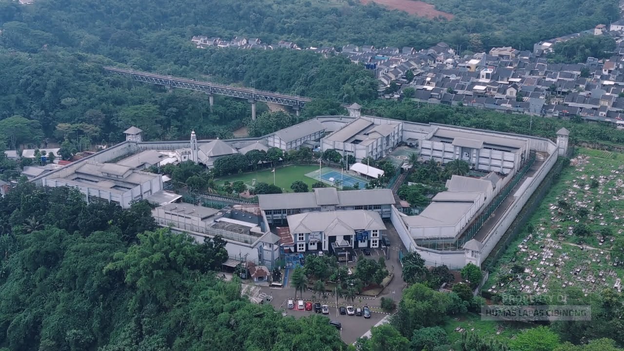 Aerial view of Cibinong Prison in Bogor, near Jakarta, surrounded by trees and a cemetery, with a parking lot and buildings visible.