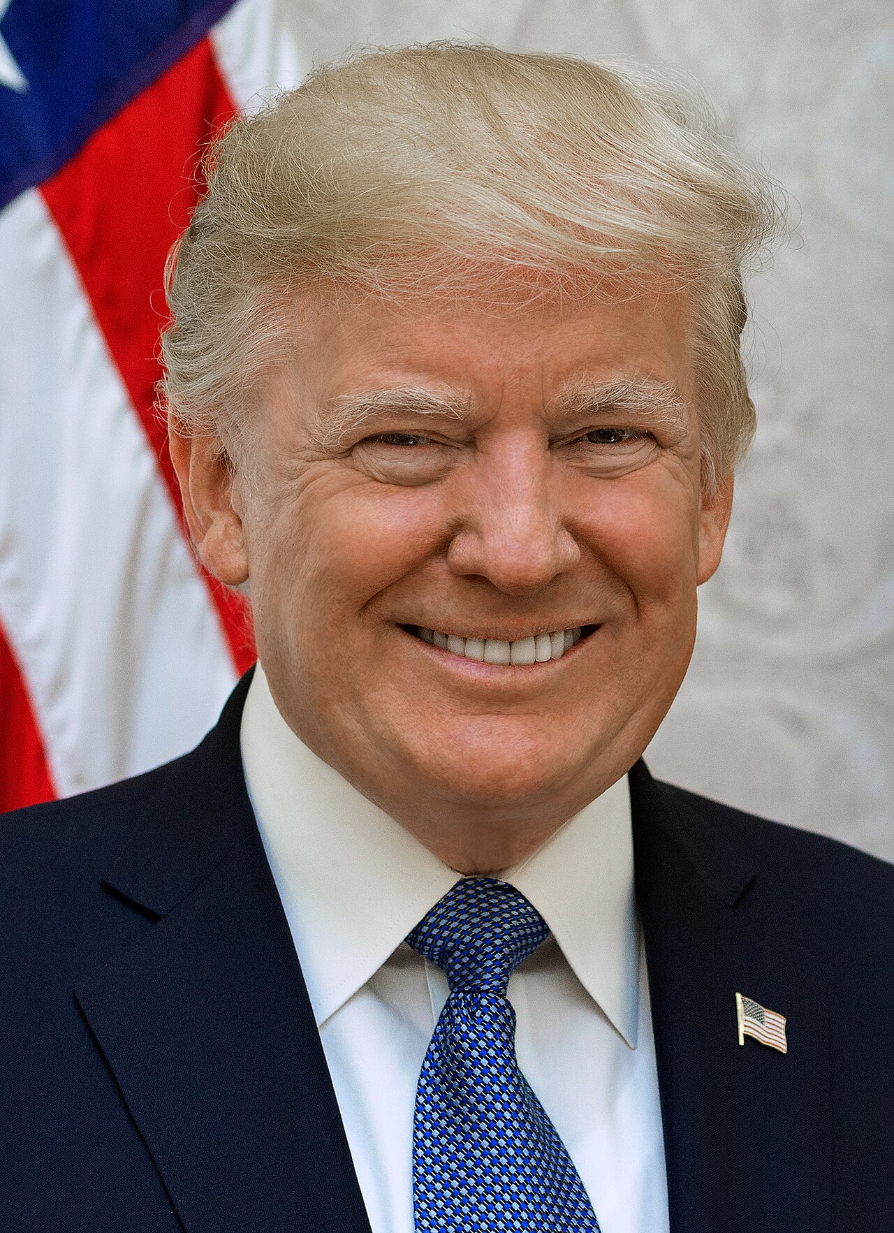 Donald Trump, a white man with blond hair, wearing a navy suit, white shirt, blue tie, and American flag pin, smiling in front of a white and American flag background.
