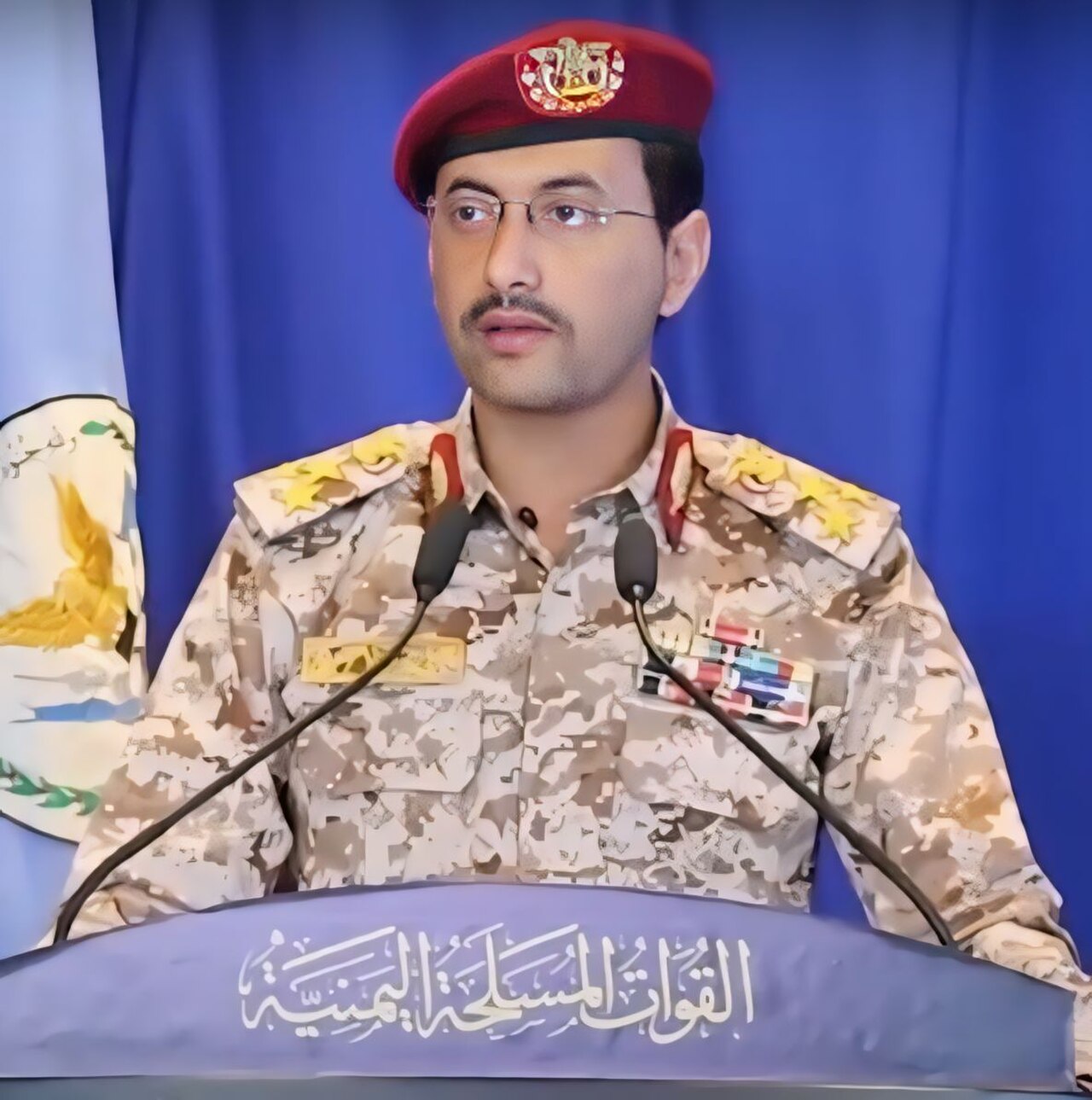 A man in a military uniform and a red beret stands behind a podium with Arabic writing, speaking into microphones.