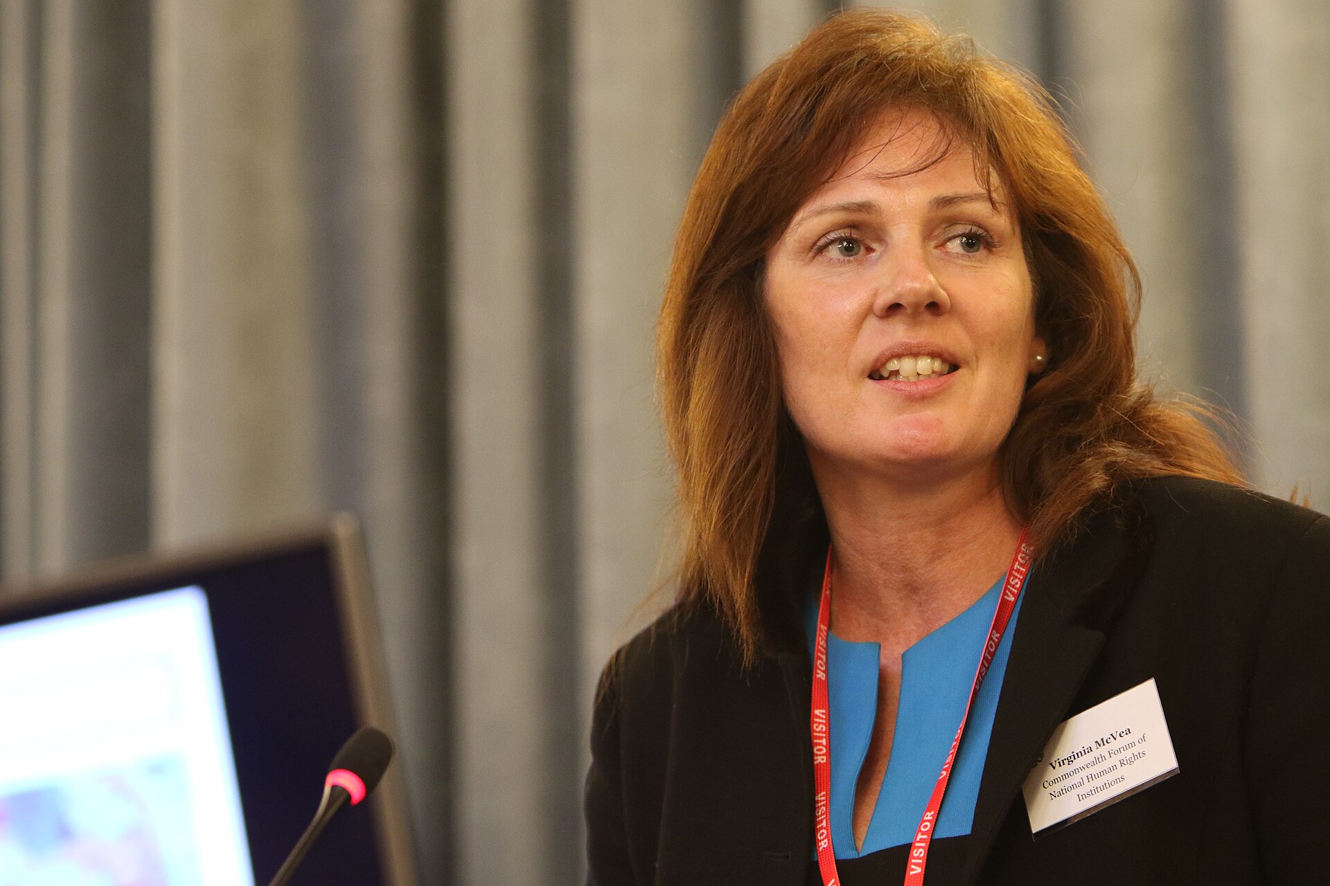 Woman with red hair wearing a black blazer, blue top, and red lanyard with a name tag that reads Virginia McVea