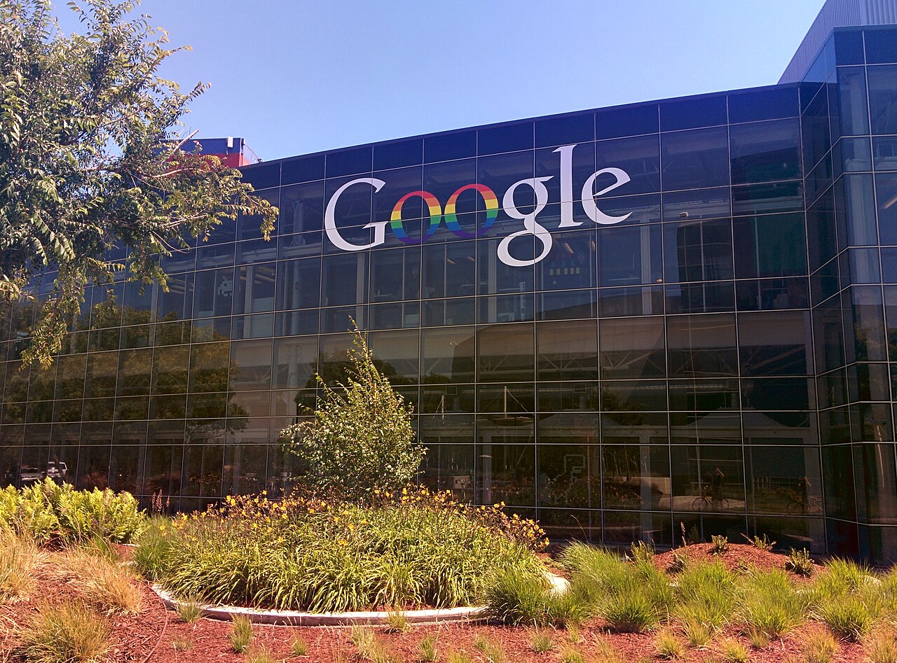 The Google logo with a rainbow-colored infinity symbol in place of the 'o's, displayed on the exterior of a modern glass building.
