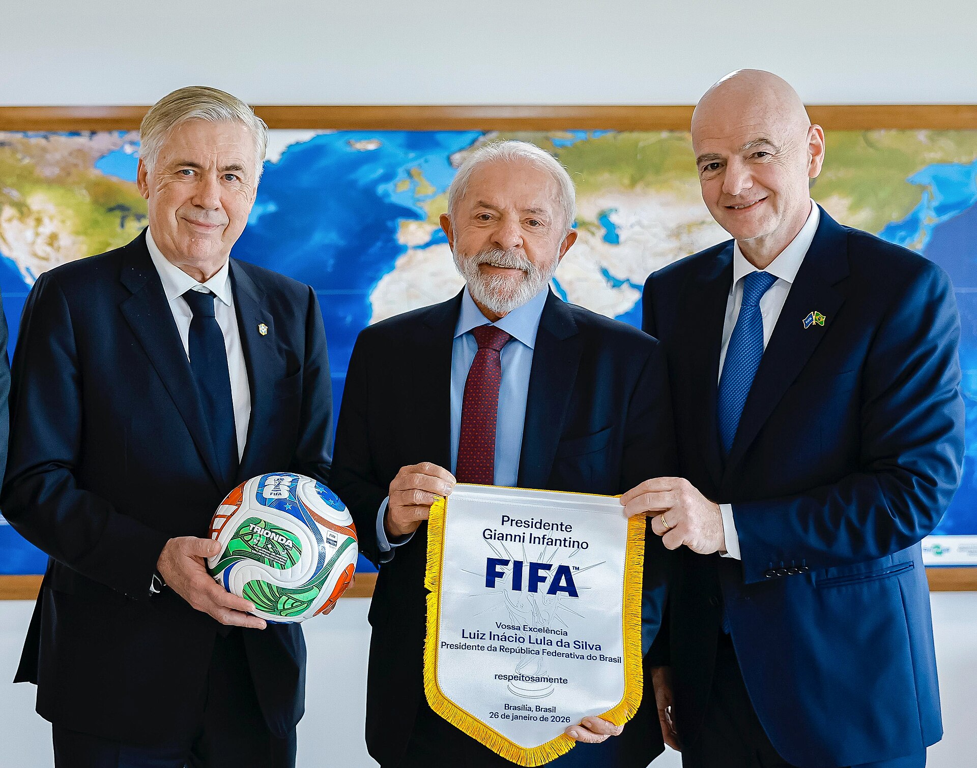 Three men in suits stand in front of a world map. The man on the right, Gianni Infantino, holds a FIFA banner. The man in the middle, Luiz Inácio Lula da Silva, holds a soccer ball. The man on the left is Carlo Ancelotti.