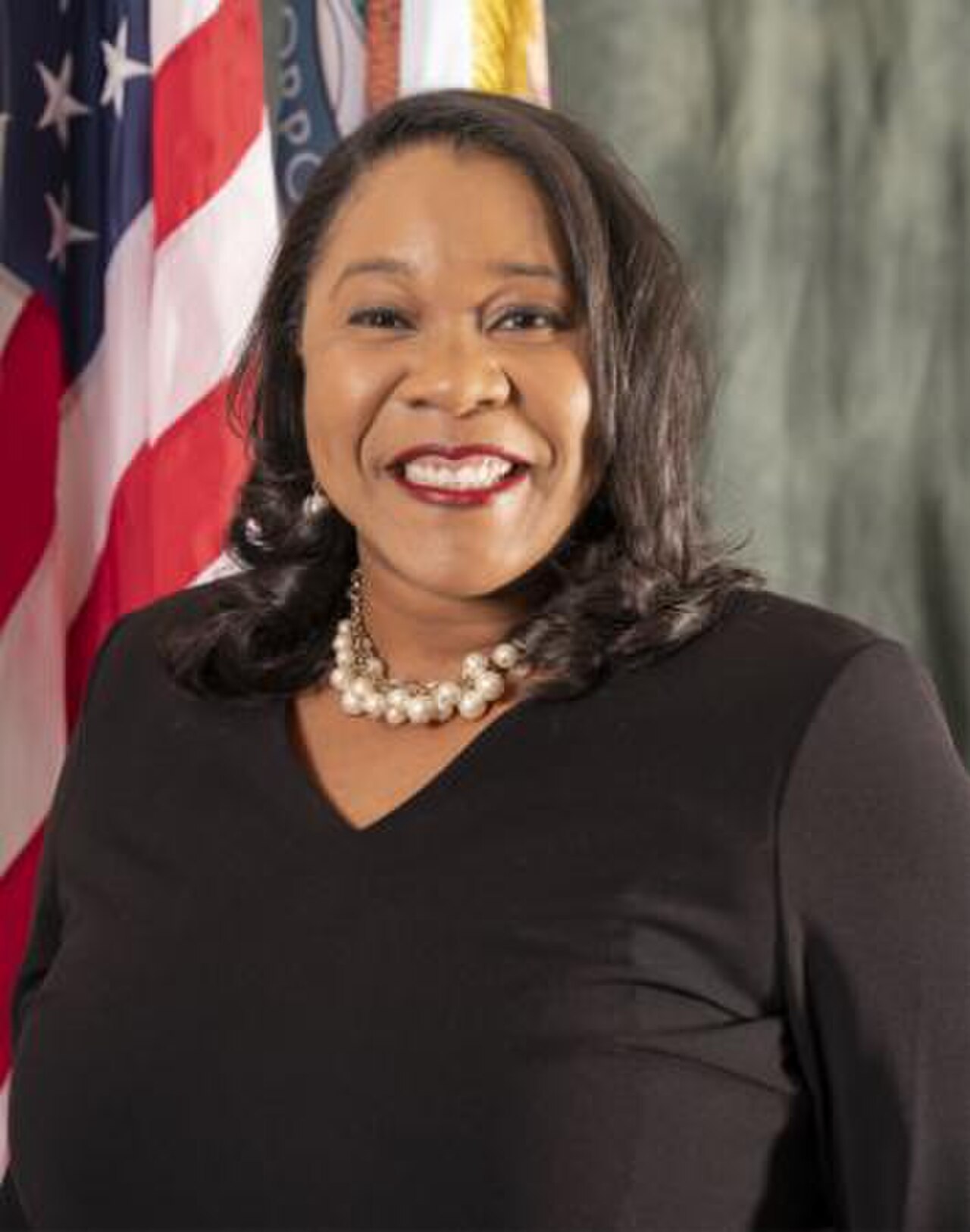 A smiling woman with dark skin and shoulder-length dark hair, wearing a black top and pearl necklace, standing in front of an American flag.