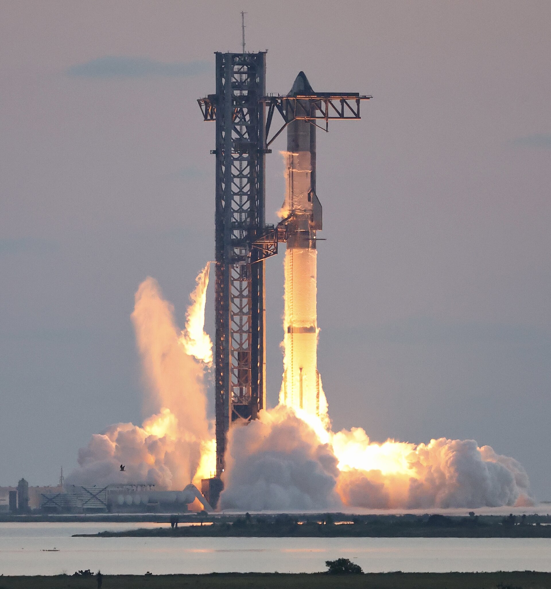 A SpaceX Starship rocket launches from a launch tower, with intense flames and smoke billowing from its base.