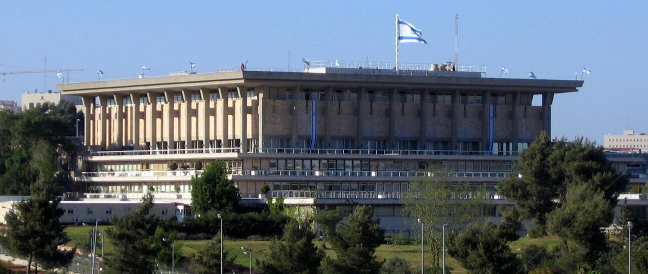 The Knesset building with Israeli flag flying, surrounded by trees and greenery