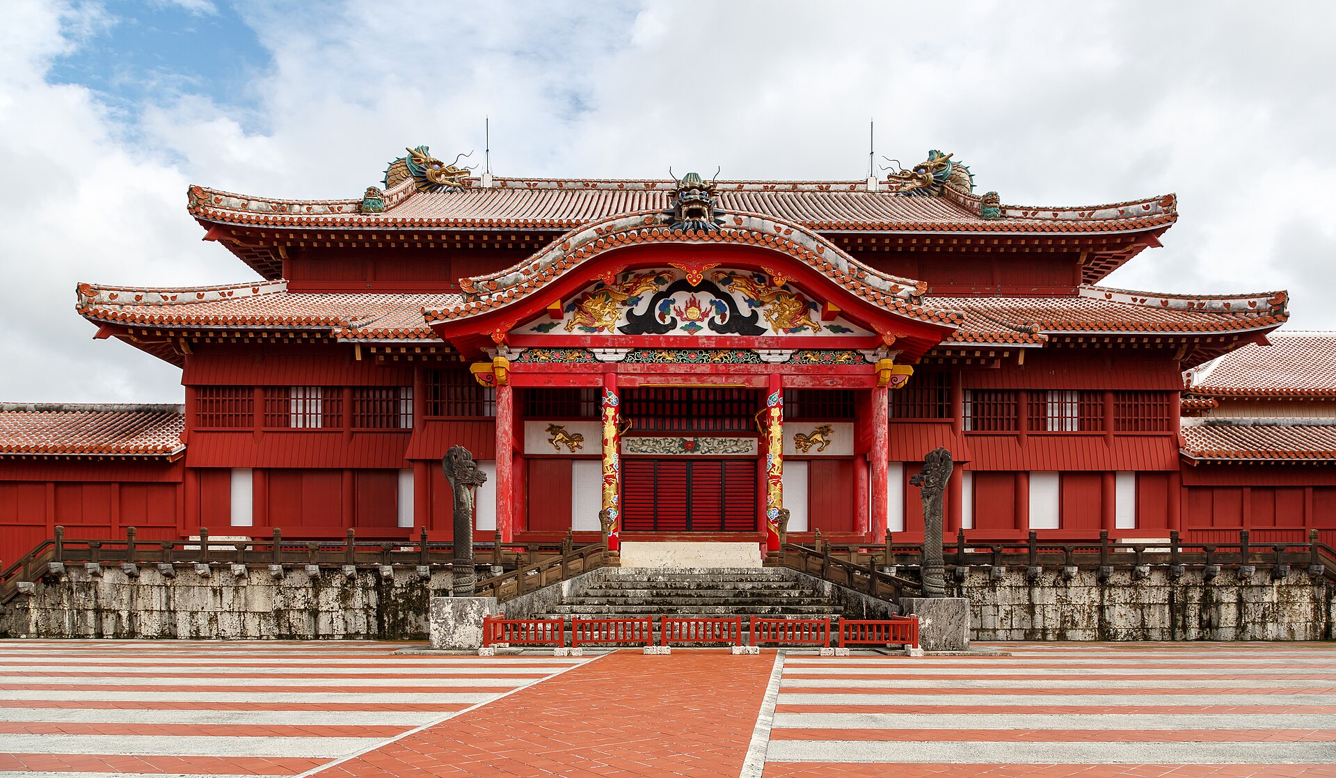 A wide shot of the Shuri Castle in Okinawa, Japan, under a cloudy sky. The castle is painted a vibrant red and features traditional Ryukyuan architecture with ornate roofs and dragon decorations. A red and white striped pathway leads up to the main entrance.