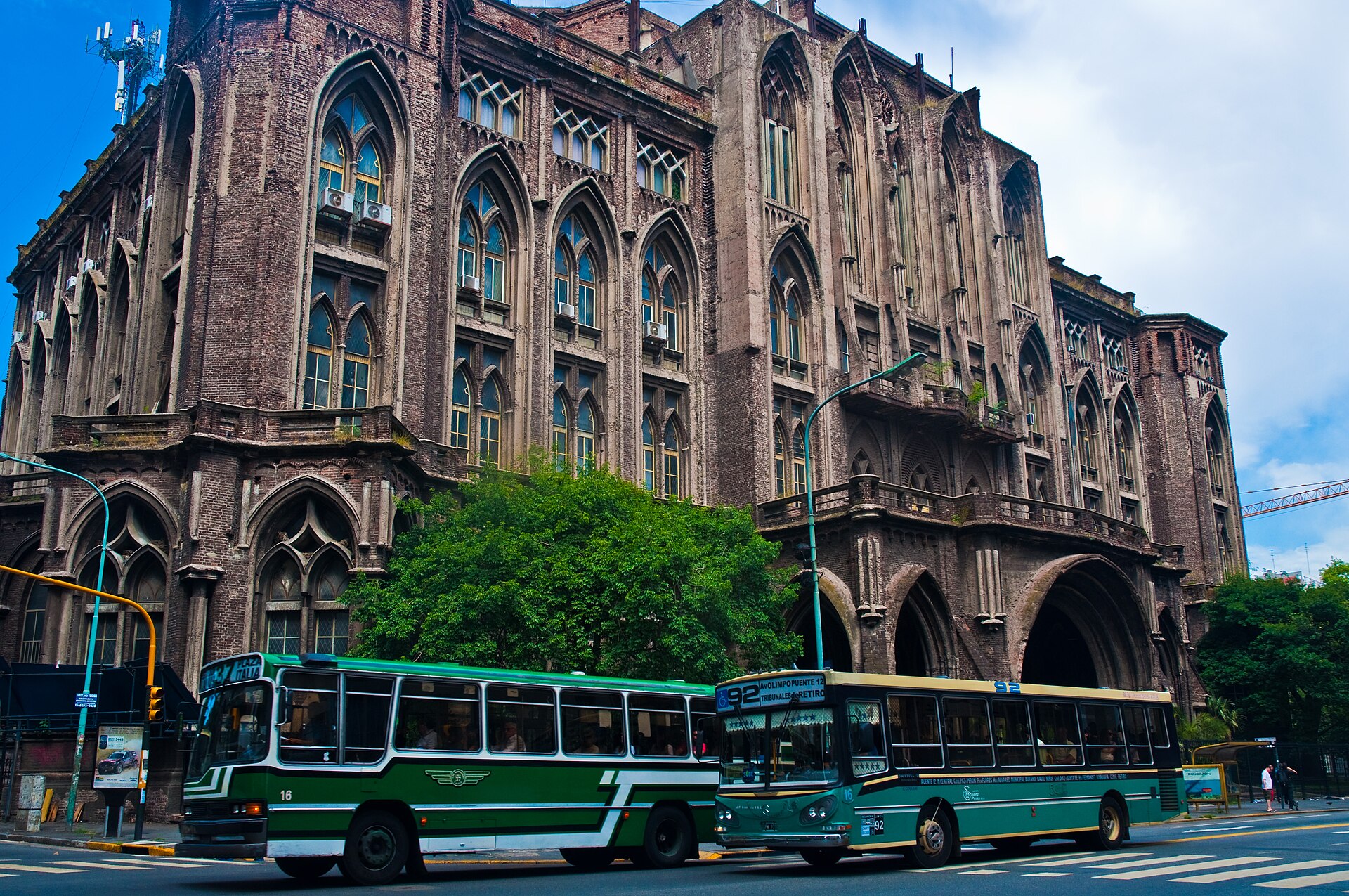 A large, ornate brick building with arched windows and balconies, with two green buses in the foreground on a street.