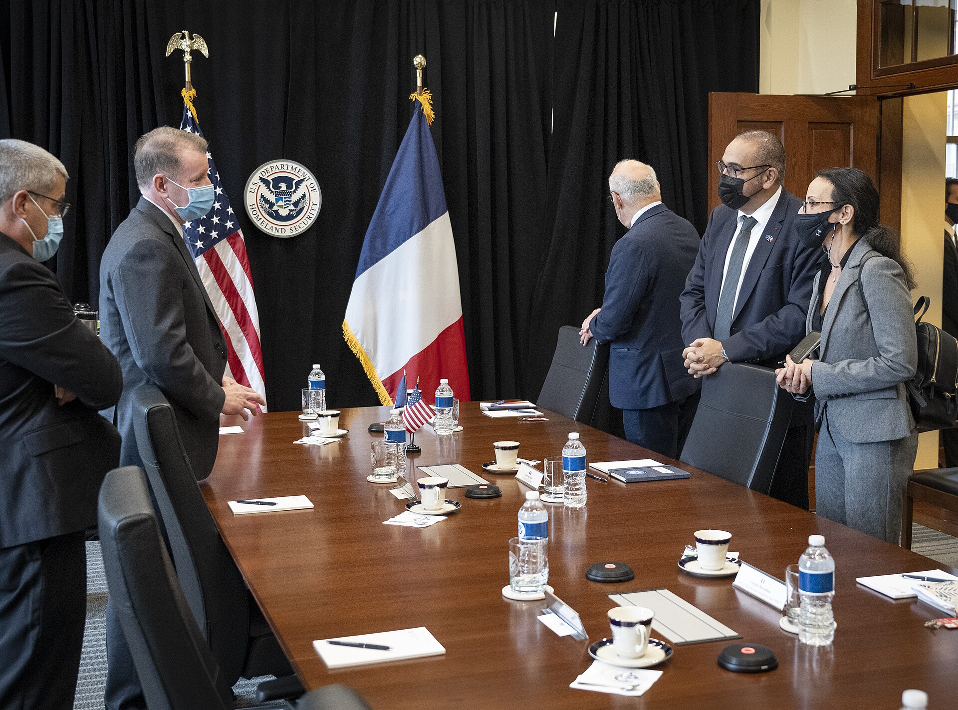 A group of people in business attire stand around a conference table, with the flags of the United States and France visible behind them. The US flag is next to a seal that reads "U.S. Department of Homeland Security".