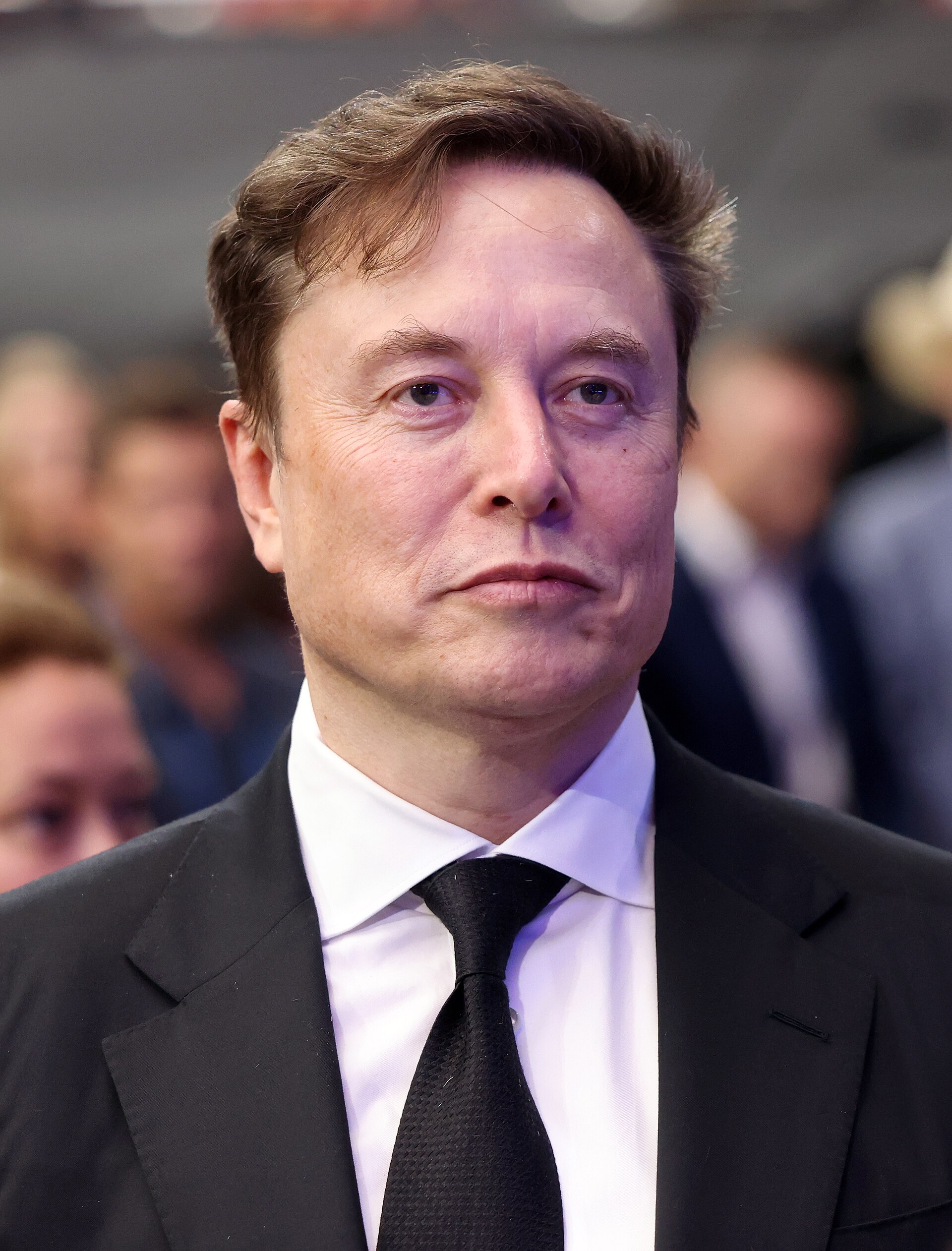 Elon Musk, wearing a black suit, white shirt, and black tie, looking to the right with a neutral expression, in front of a blurred background of people