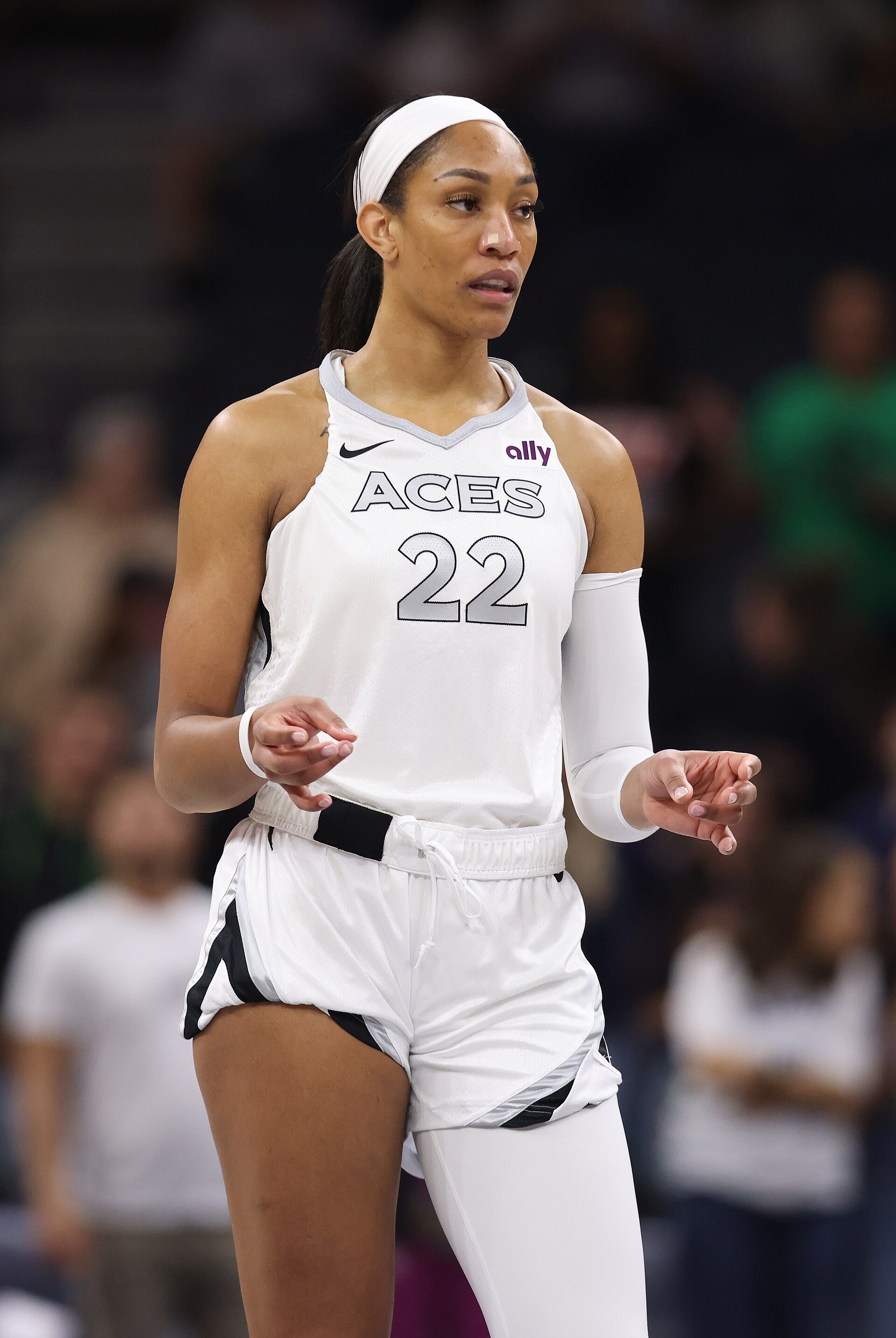 A'ja Wilson, a black woman with dark hair pulled back, wearing a white Aces 22 jersey, white arm sleeve, white shorts, and white leggings, standing on a basketball court with her hands outstretched.