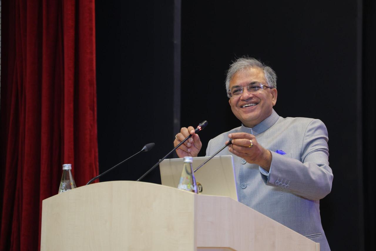 A man with gray hair and glasses in a light blue suit speaking at a podium with two microphones and two water bottles.