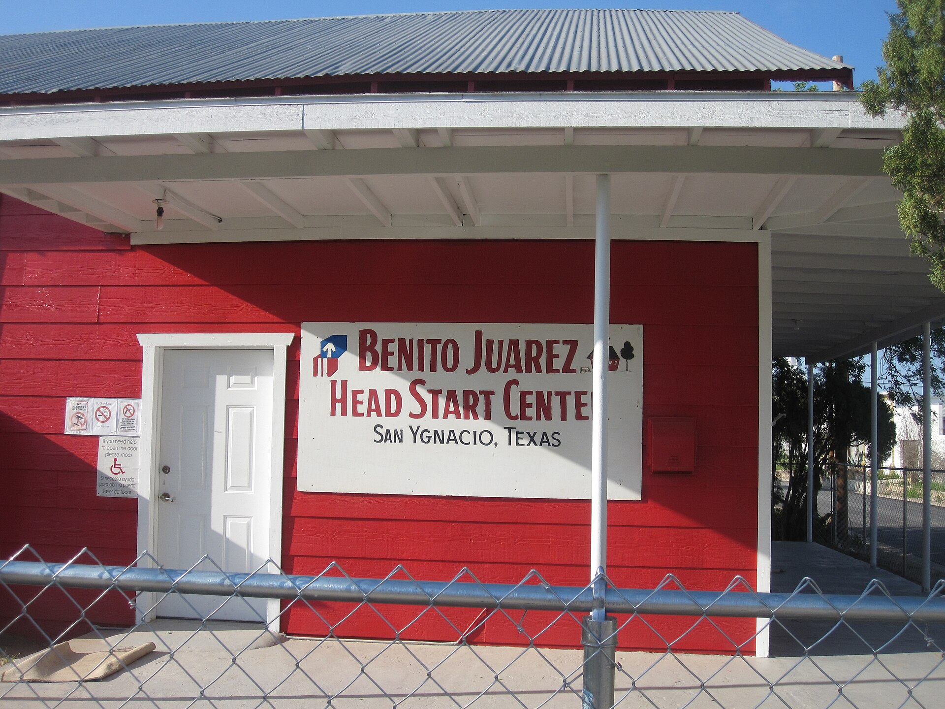 A red building with a sign that reads "BENITO JUAREZ HEAD START CENTER SAN YGNACIO, TEXAS". There is a white door to the left of the sign and a chain-link fence in the foreground.