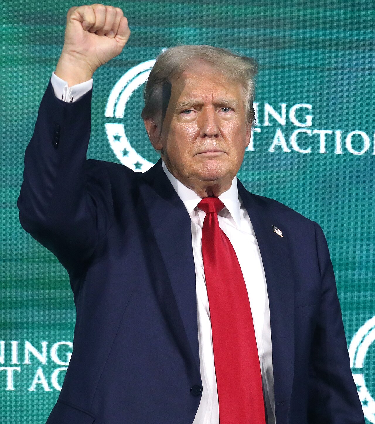 Donald Trump raises his fist in the air while wearing a dark suit, white shirt, and red tie. A green background with white text is visible behind him.