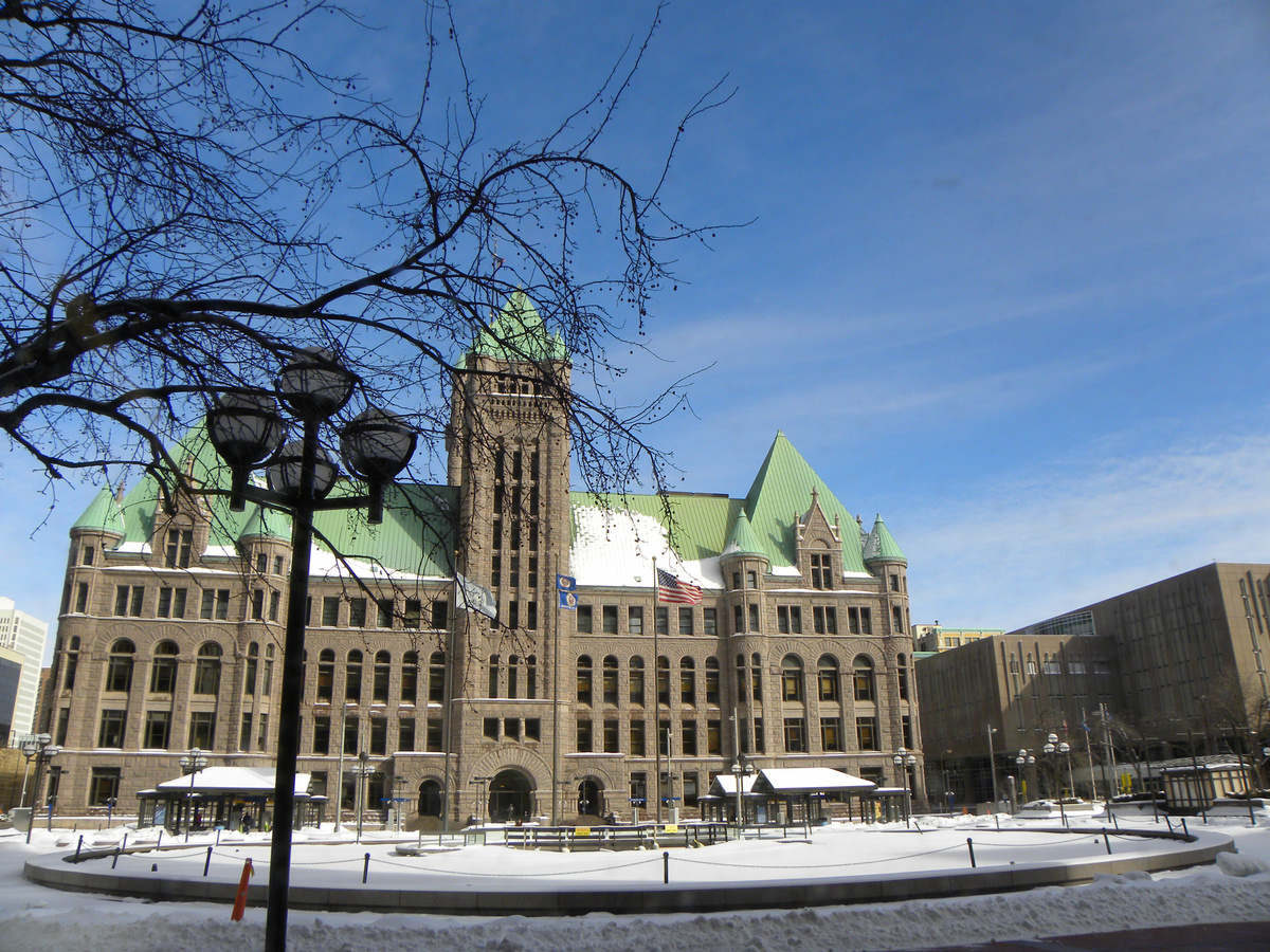 Minneapolis City Hall in winter, a stone building with a green copper roof and clock tower, snow on the plaza in front.