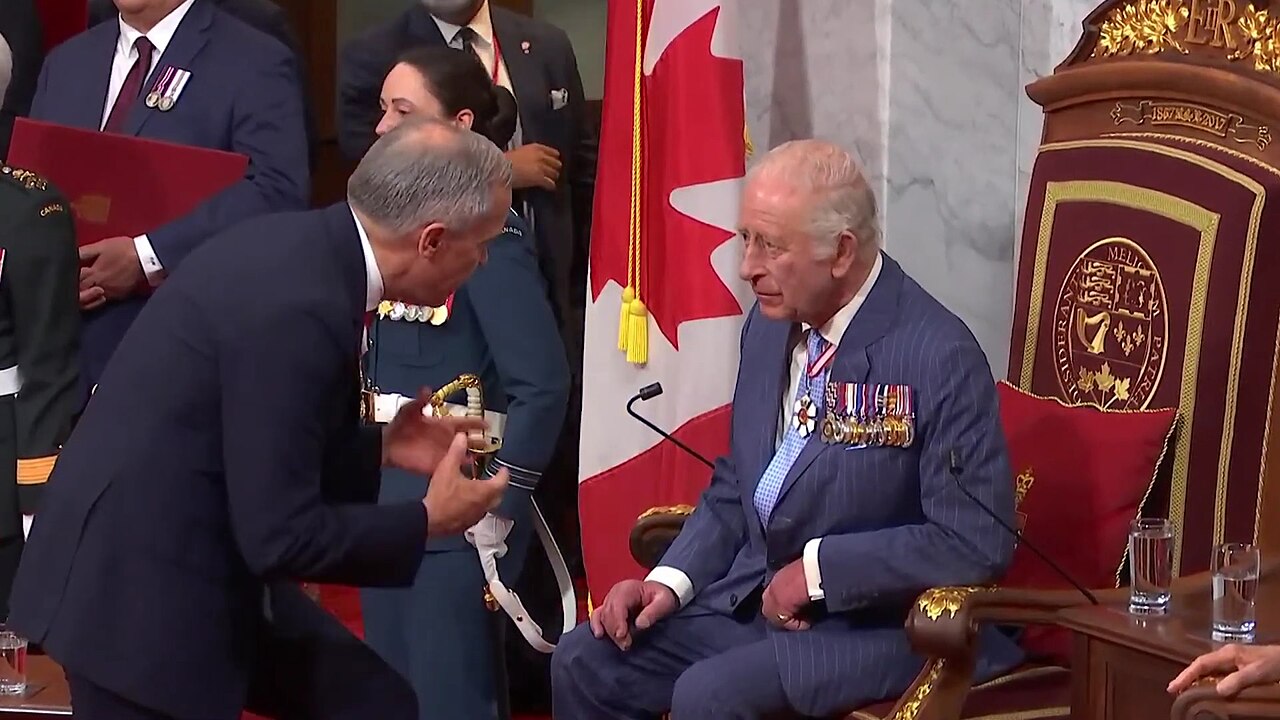 King Charles III seated, wearing a blue suit with medals, and a man in a dark suit bowing to him, with a Canadian flag and a military uniform in the background.