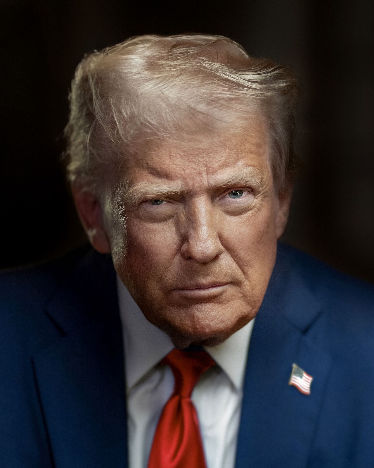 Donald Trump, wearing a blue suit, white shirt, red tie, and American flag pin, looking down