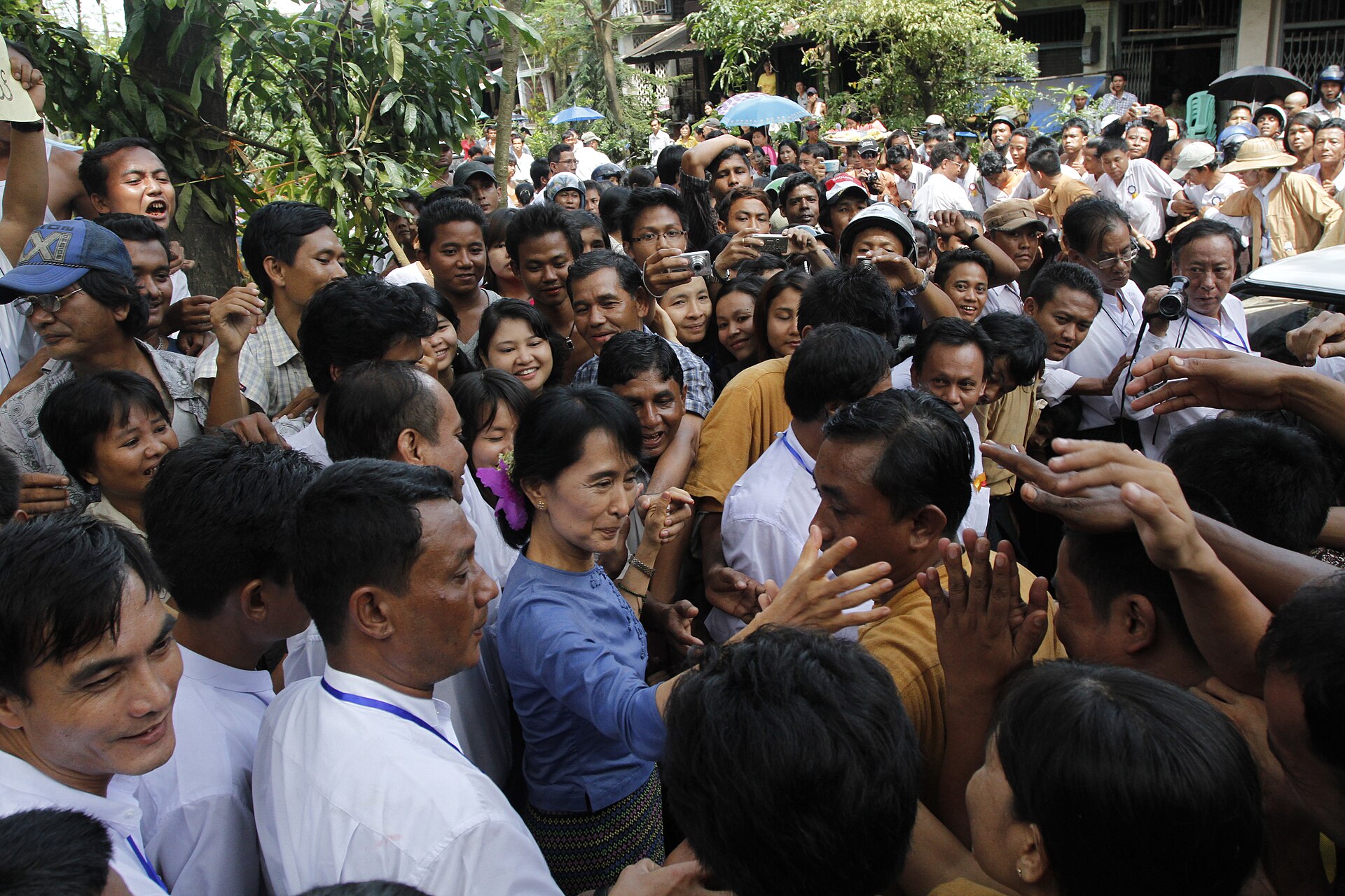Aung San Suu Kyi, smiling, in a blue shirt and patterned skirt, is surrounded by a crowd of people, some of whom are reaching out to her.