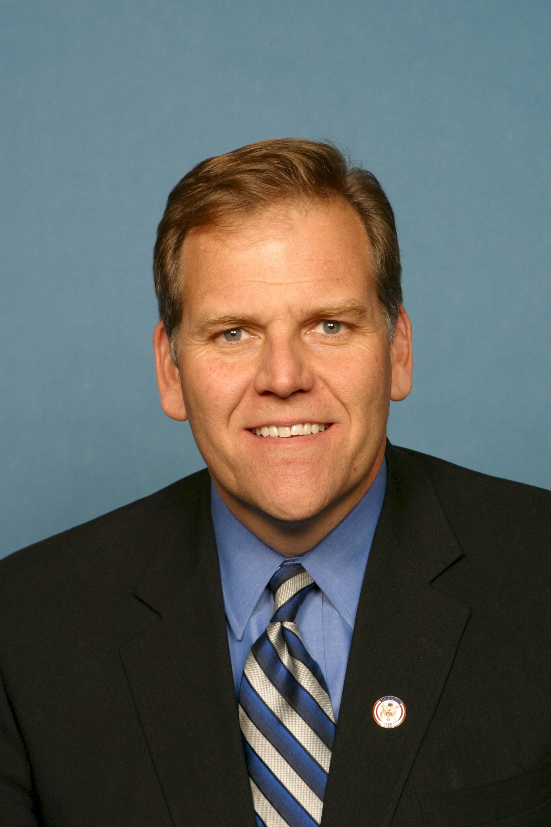 A headshot of a white man with short brown hair, blue eyes, and a slight smile, wearing a dark suit, a light blue collared shirt, and a striped tie. A circular pin with an eagle emblem is visible on his left lapel.