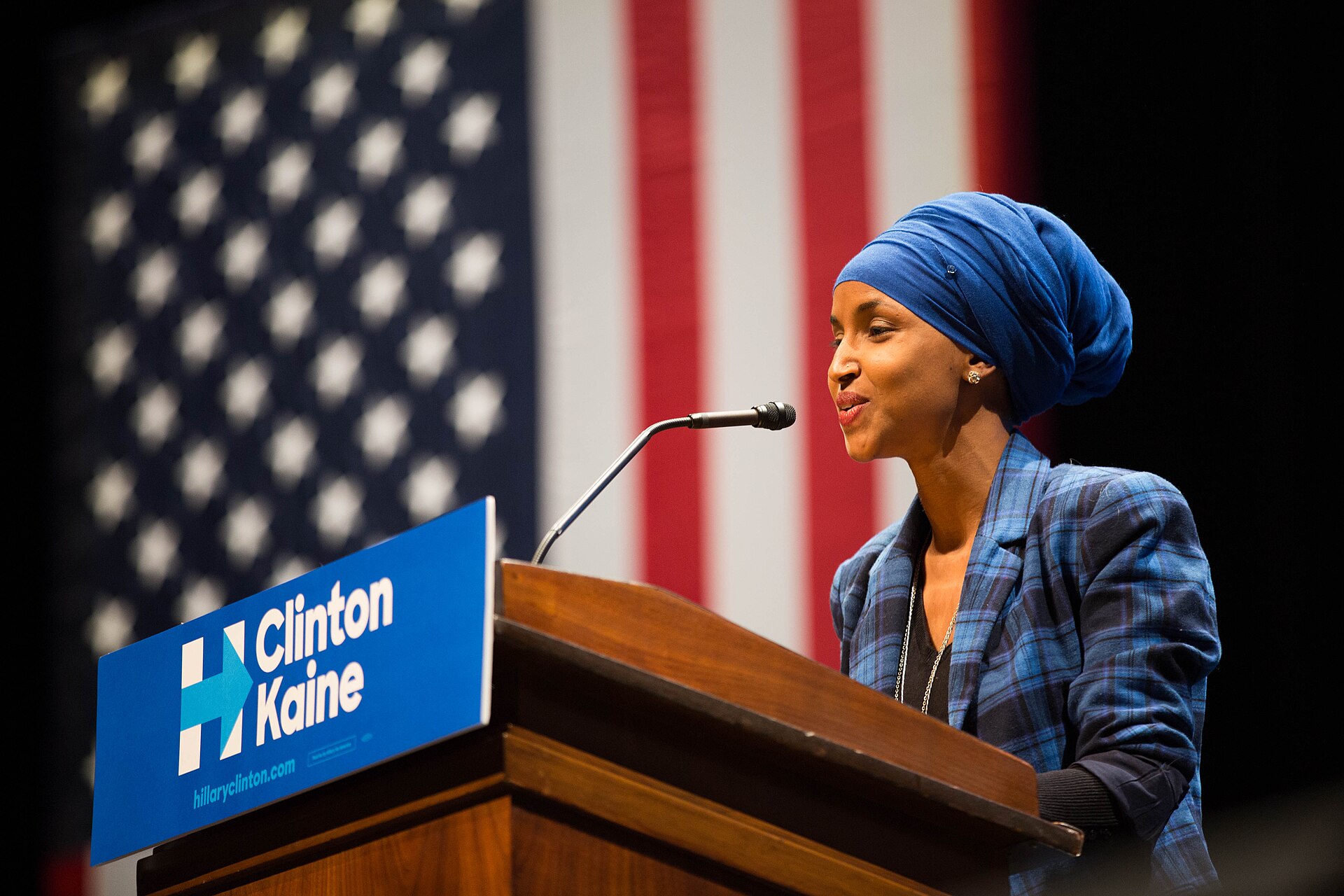 A woman wearing a blue headscarf and a blue plaid jacket speaks at a podium with a "Clinton Kaine" sign in front of an American flag.