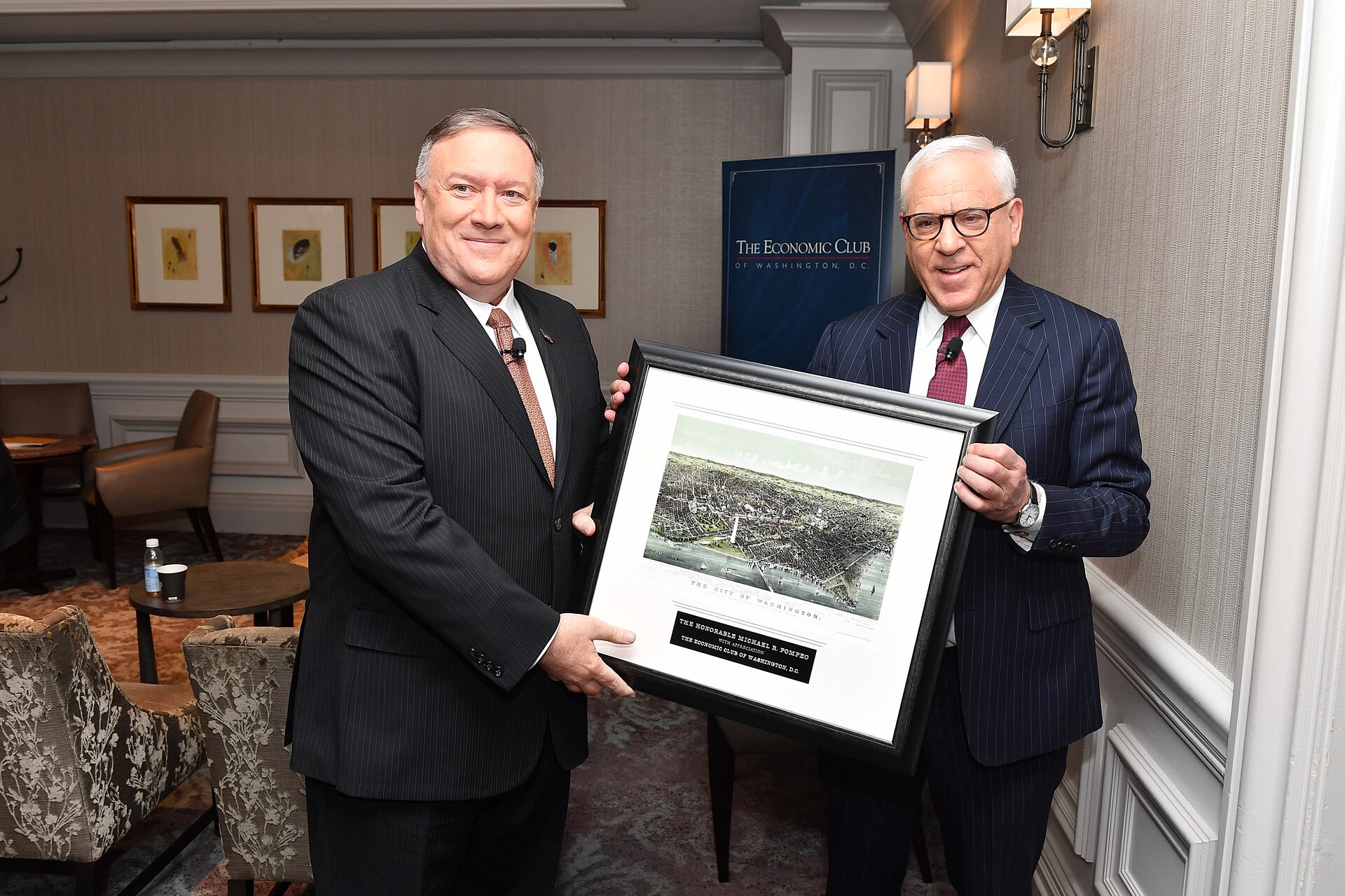 Two men in suits stand holding a framed picture of a city. The man on the left is wearing a pinstripe suit and a red tie. The man on the right is wearing a dark pinstripe suit and a red tie. Behind them is a blue banner with "The Economic Club of Washington, D.C." written on it.