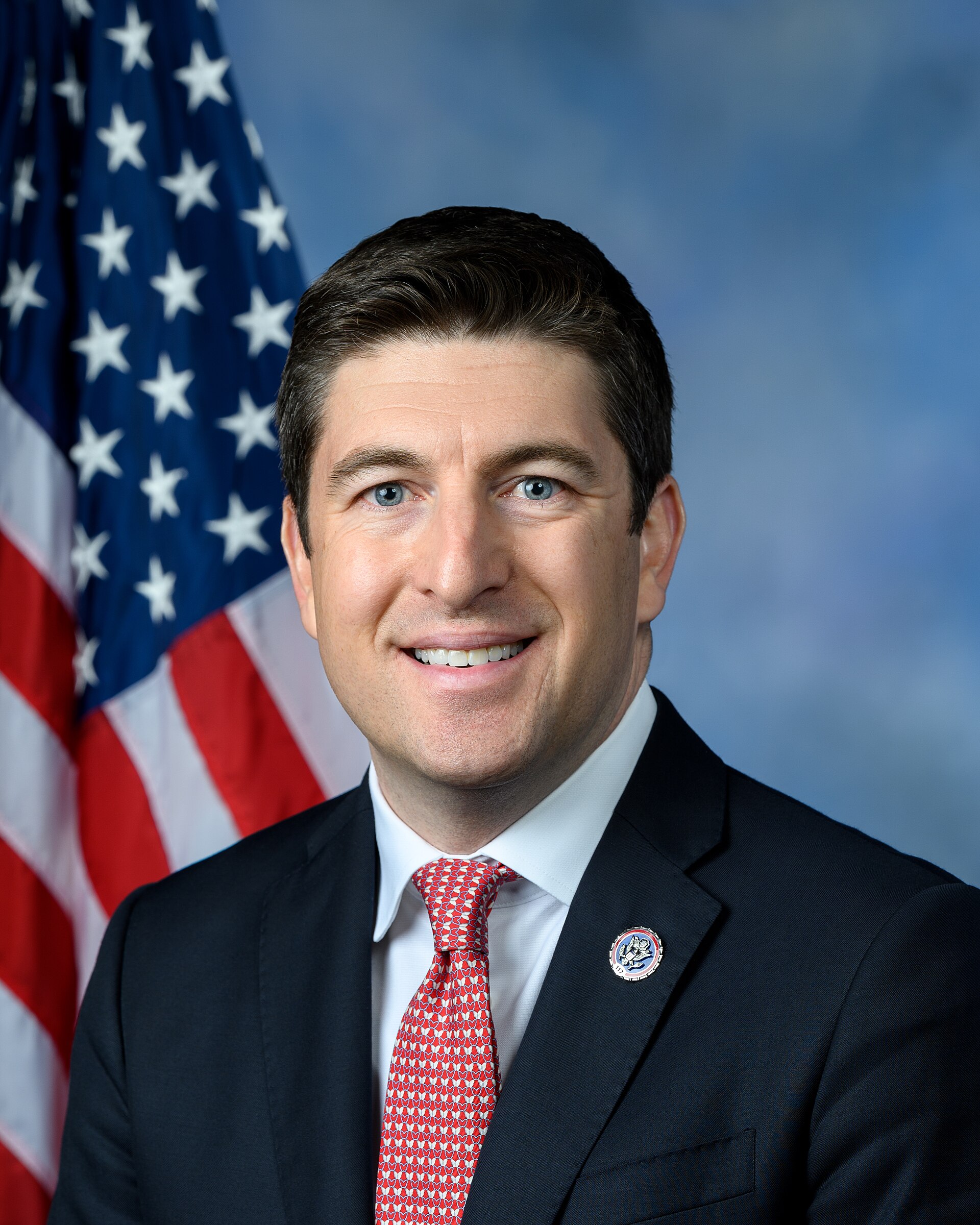 Bryan Steil, a middle-aged man with short dark hair, blue eyes, and a slight smile, wearing a navy suit, white shirt, and red tie with white dots, with an American flag pin on his lapel, standing in front of an American flag and a blue background.