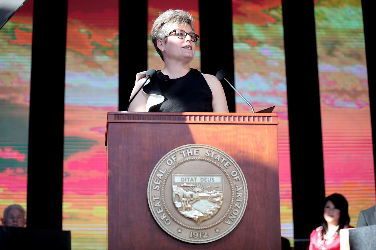 A woman with short gray hair and glasses stands at a podium with two microphones and the seal of the state of Arizona.