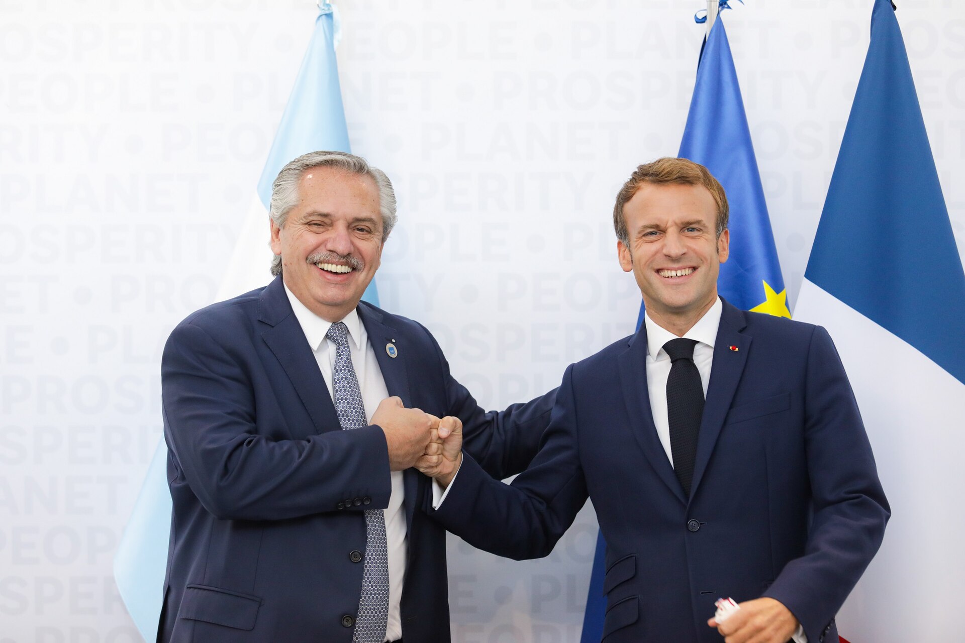 Two men in suits are bumping fists in front of flags.