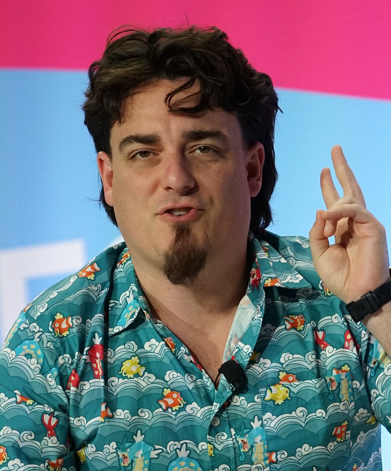 A man with dark hair, a goatee, and a blue shirt with a fish pattern, gesturing with his hand.