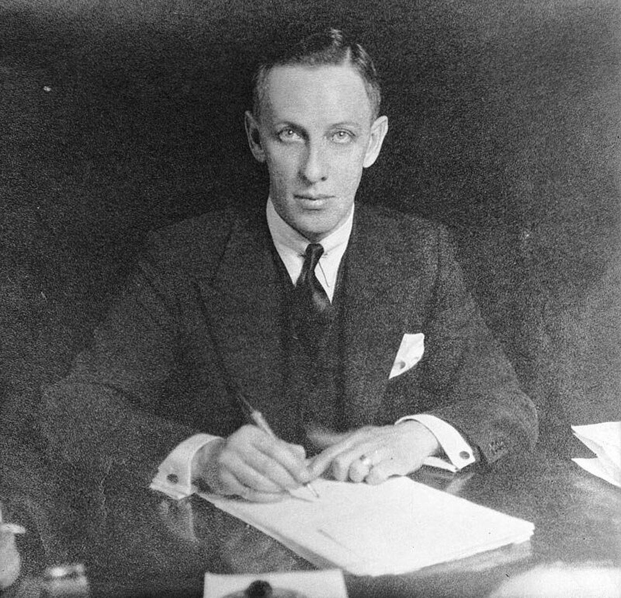 A man in a suit sitting at a desk writing on a piece of paper