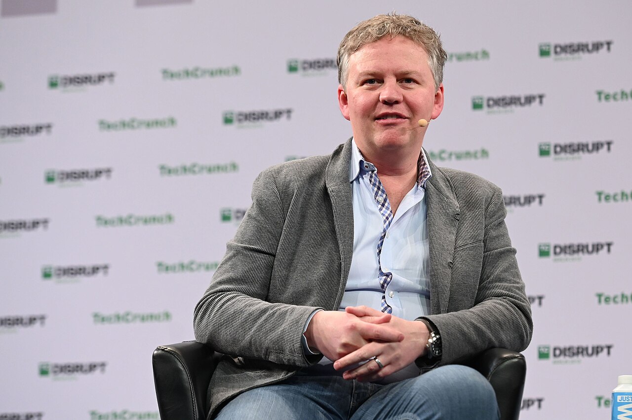 A man with short, light brown hair, wearing a grey blazer over a light blue collared shirt and jeans, sits with his hands clasped in his lap. He is wearing a ring on his left ring finger and a watch on his left wrist. The background is a white wall with repeating logos that appear to be "TechCrunch