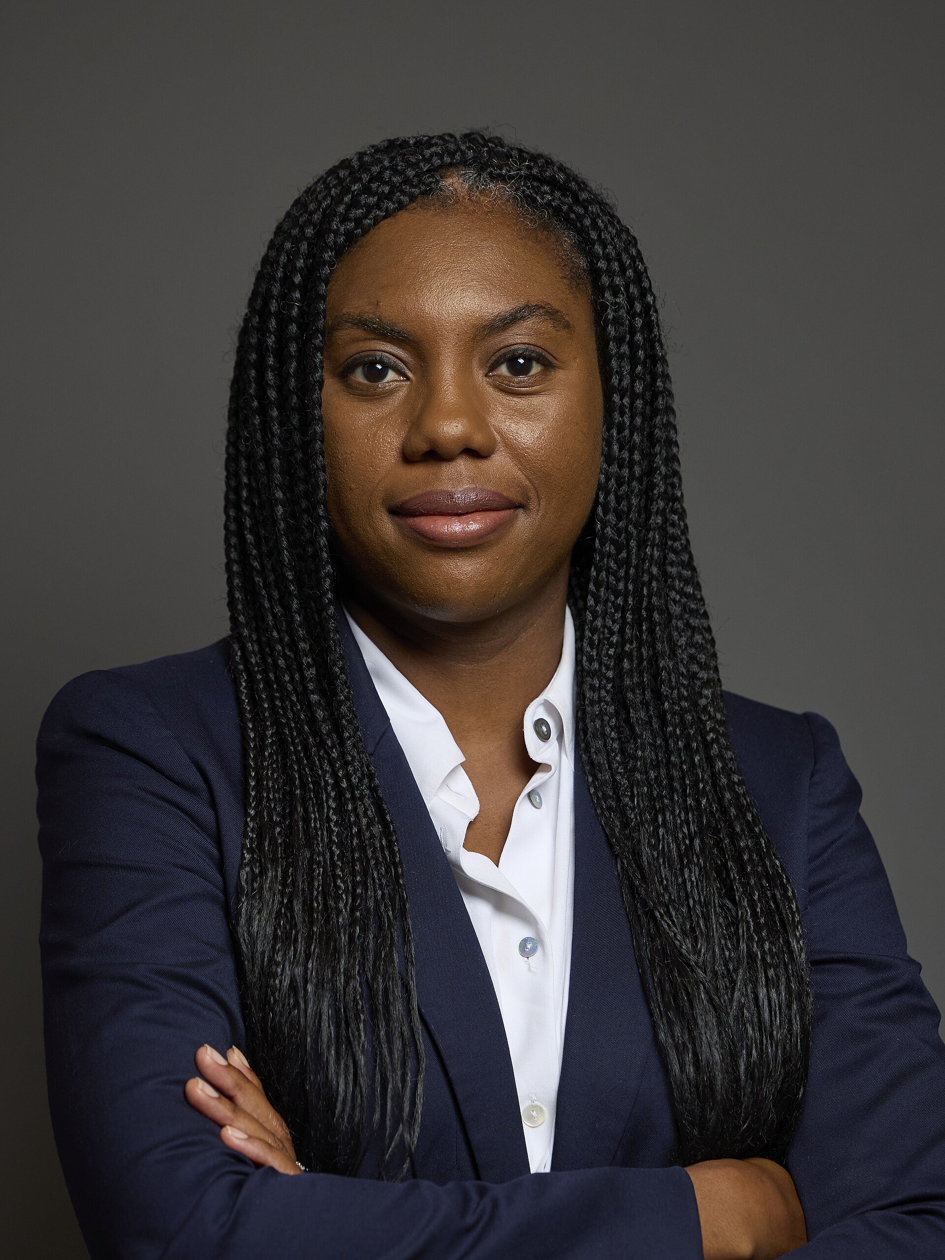 A woman with long black braids wearing a white collared shirt and a dark blue blazer, with her arms crossed, against a gray background.
