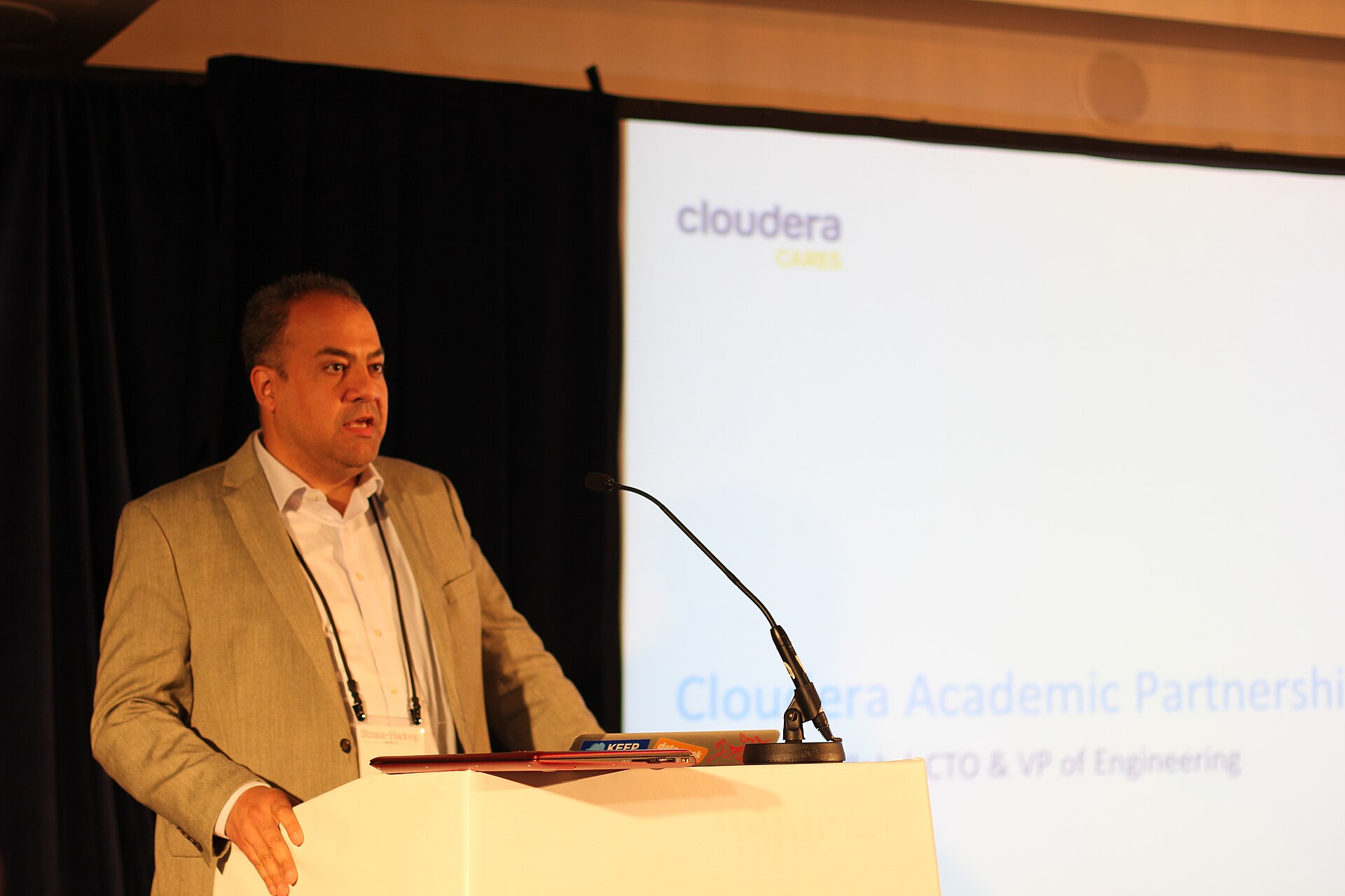 A man in a suit stands at a podium with a microphone in front of a screen displaying the Cloudera logo and text that reads "Cloudera Academic Partnership" and "CTO & VP of Engineering."