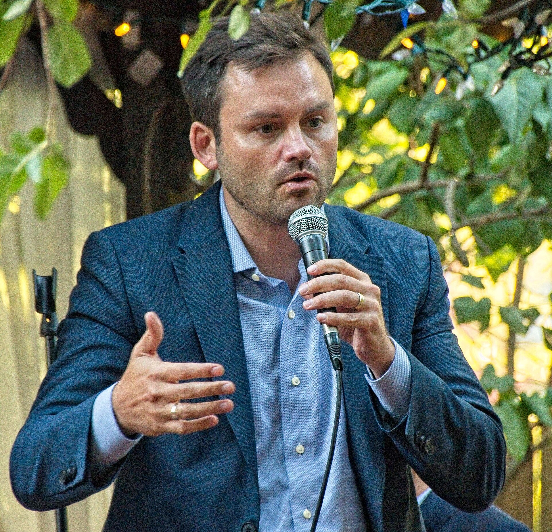 A man in a blue suit jacket and light blue shirt speaking into a microphone.