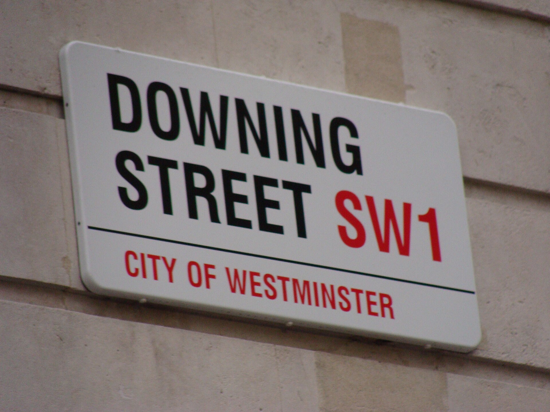 A white street sign with black text reading "DOWNING STREET SW1" and red text below reading "CITY OF WESTMINSTER".