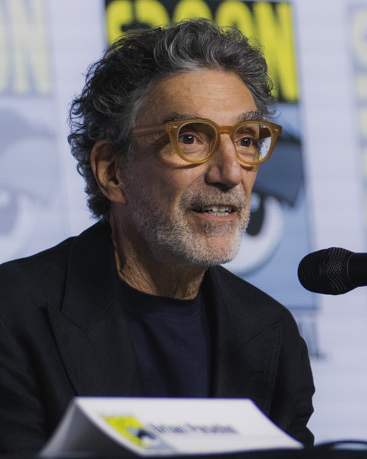 man with graying curly hair, brown eyes, and amber-framed glasses, wearing a black jacket and dark shirt, speaking into a microphone