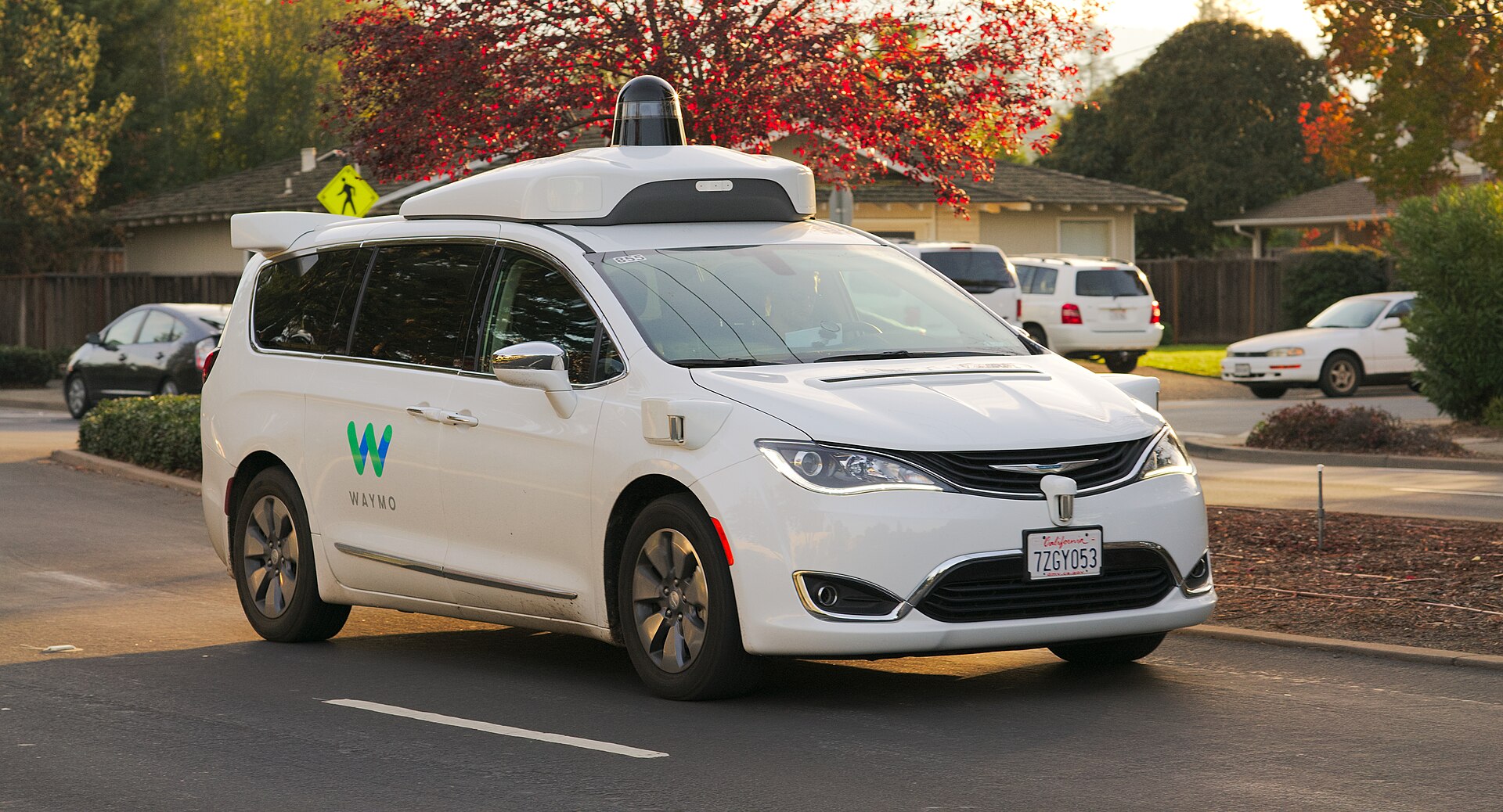 A white Waymo autonomous vehicle drives on a suburban street.