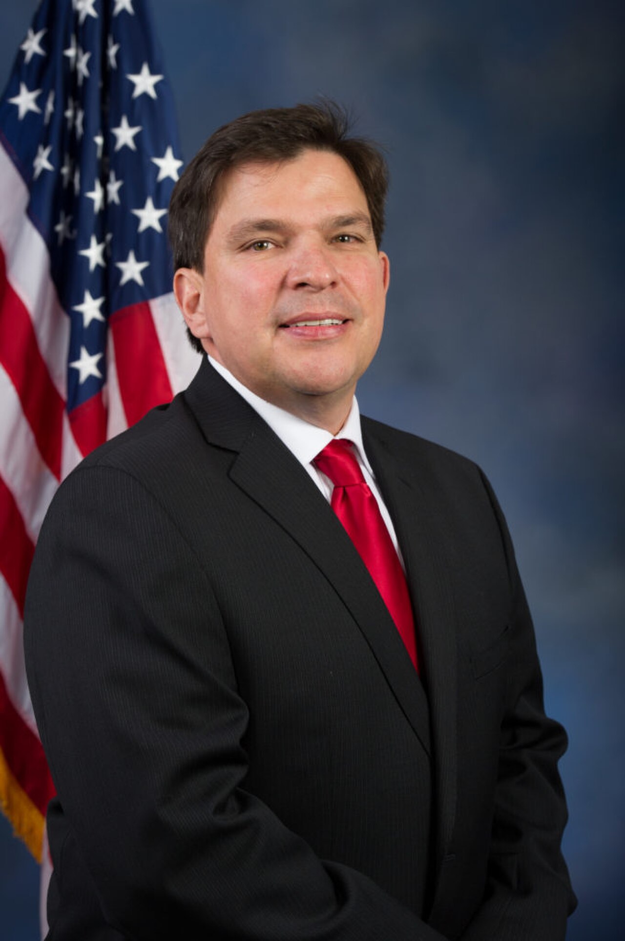 A man in a black suit, white shirt, and red tie stands in front of an American flag.