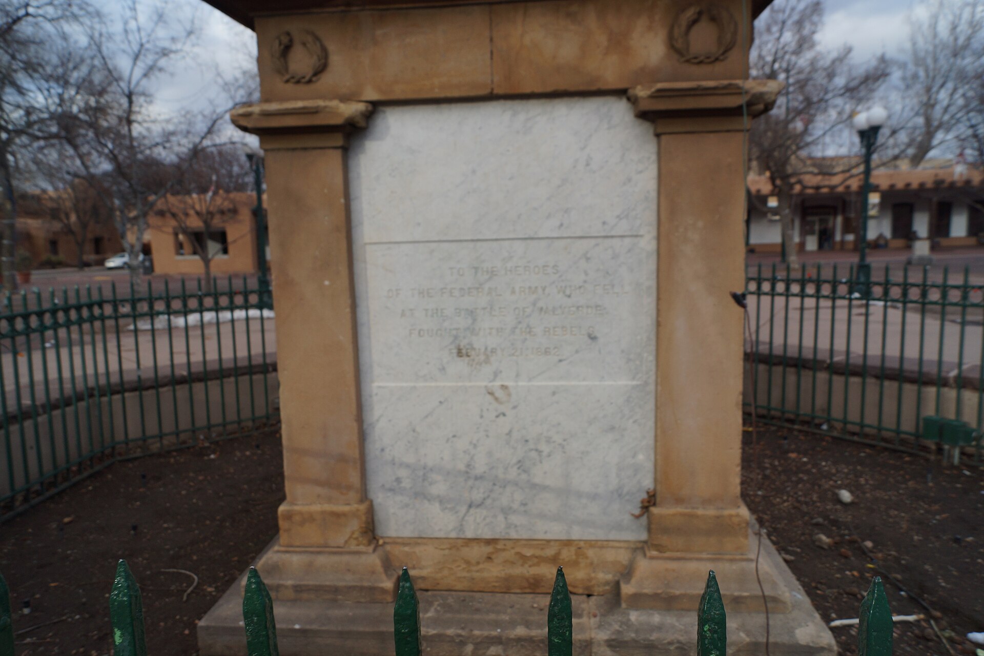 A stone monument with an inscription in the center, surrounded by a green metal fence.