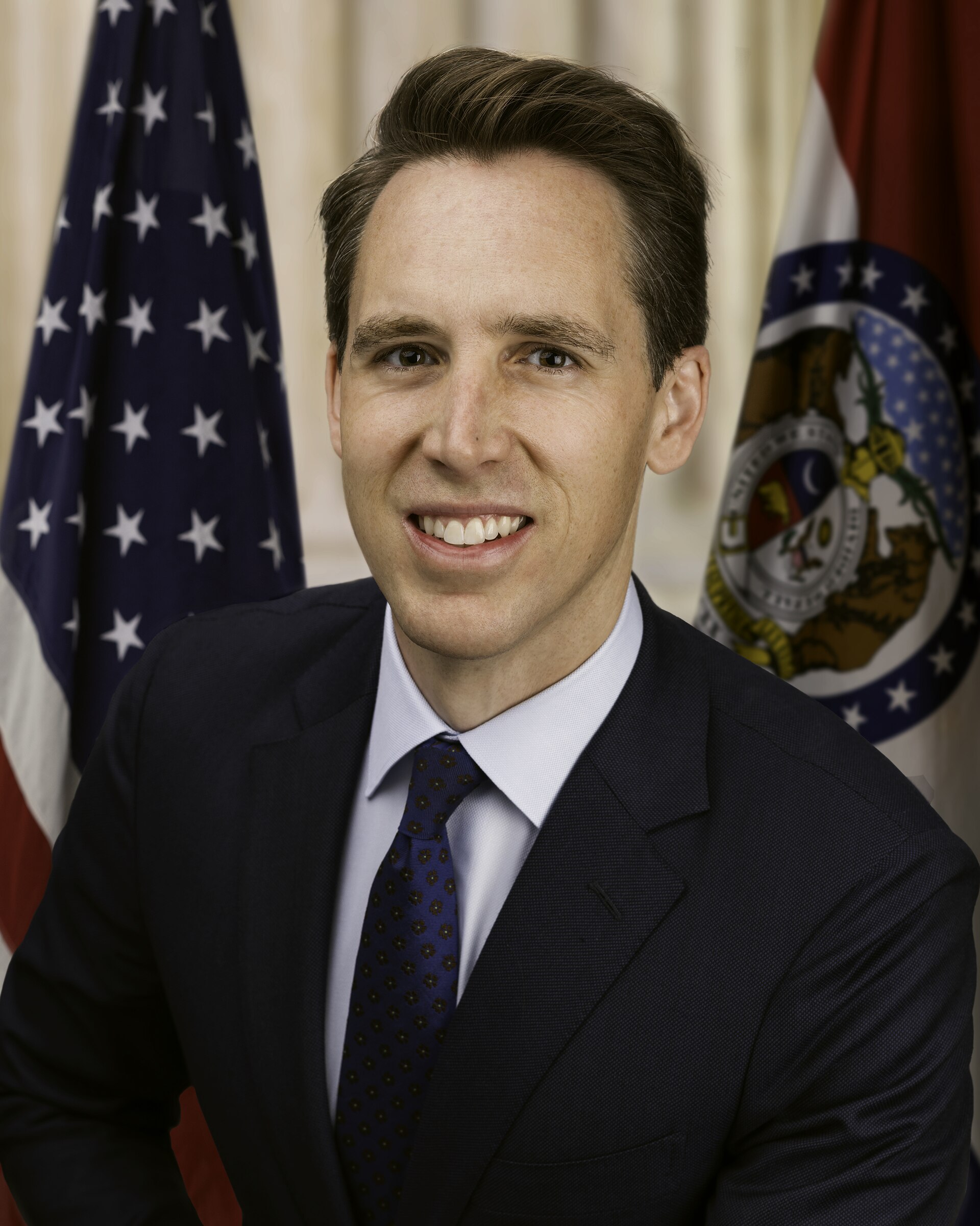 A portrait of Senator Josh Hawley, a white man with brown hair, wearing a dark suit, light blue shirt, and a blue patterned tie. He is smiling and looking at the camera. Behind him are the American flag and the Missouri state flag.