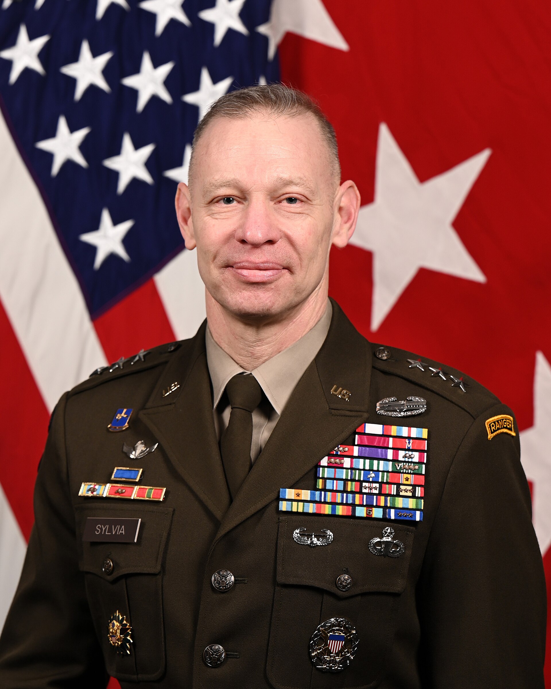 A man in a U.S. Army uniform with multiple ribbons and badges stands in front of an American flag and a red background with a white star.